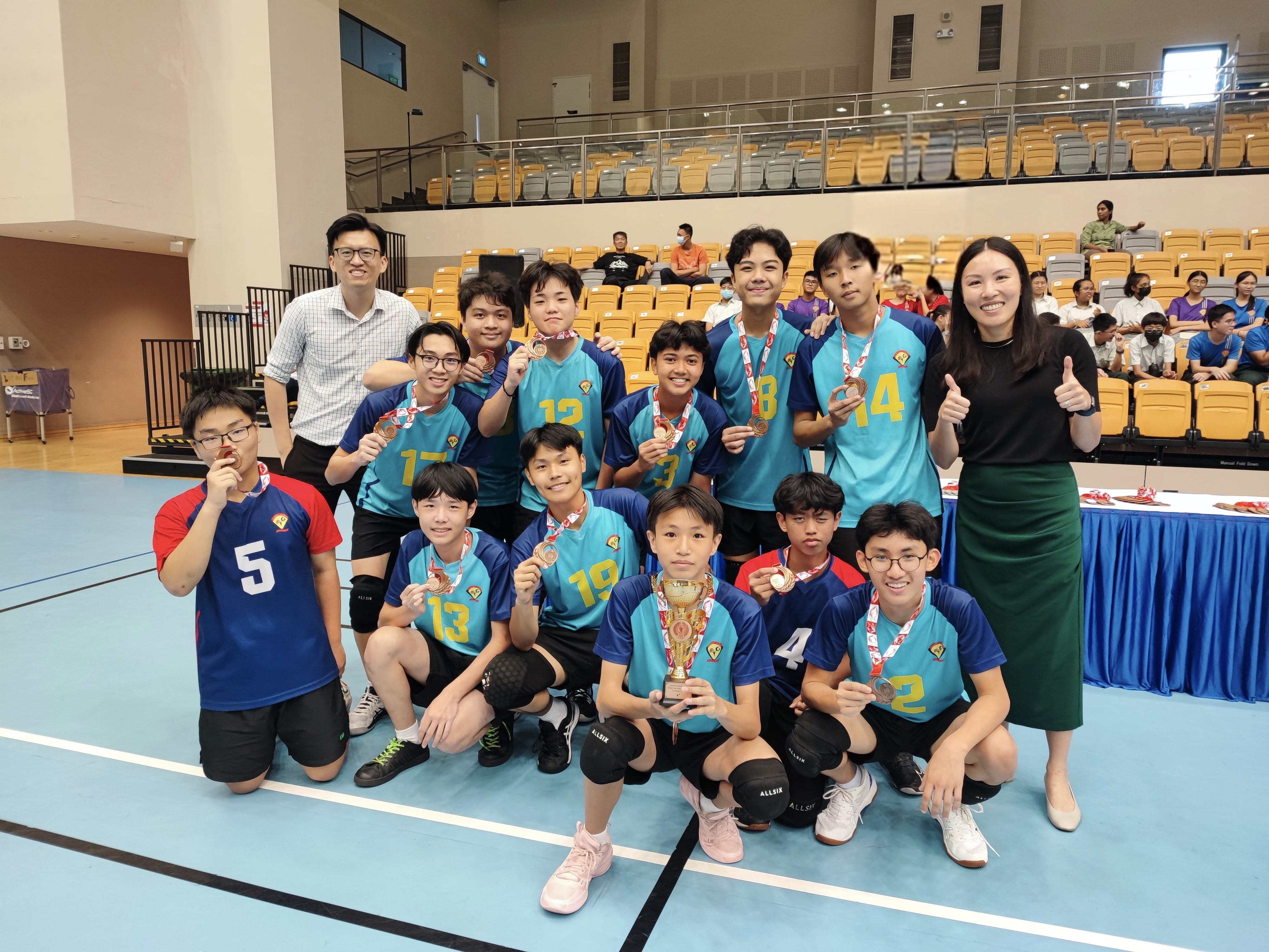 A youth volleyball team in teal jerseys poses with medals and a trophy on an indoor court, flanked by two adult coaches.