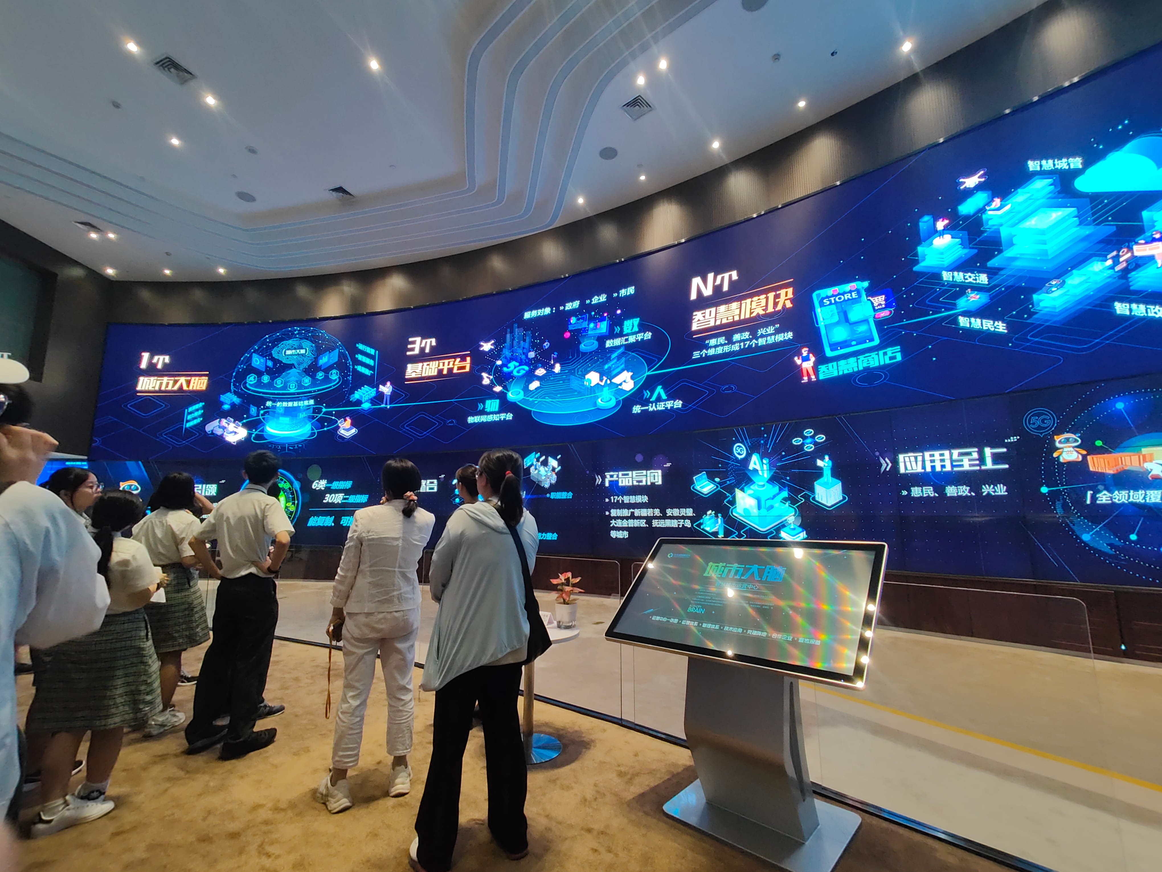 Visitors stand before a curved wall of blue digital displays with Chinese text and tech diagrams; kiosk screen at right.