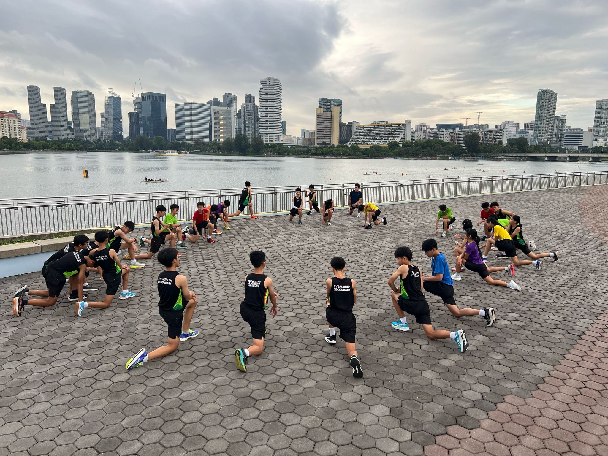 Students in Evergreen Secondary jerseys do lunges on a riverside plaza with a city skyline behind them.
