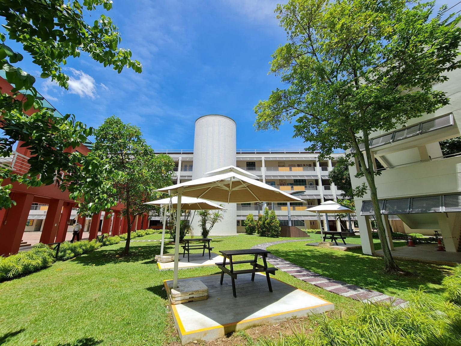 Outdoor courtyard with picnic tables under white umbrellas, green lawn, trees, and multi-story buildings under a blue sky.