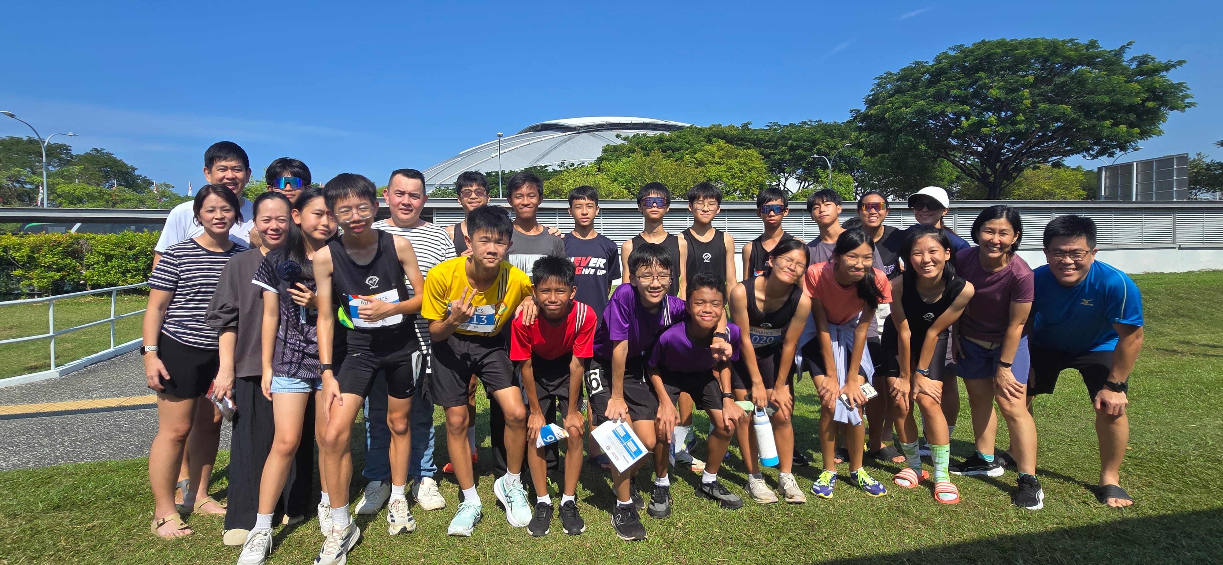 A large group of youth and adults in athletic wear pose together on grass outdoors under a clear blue sky.