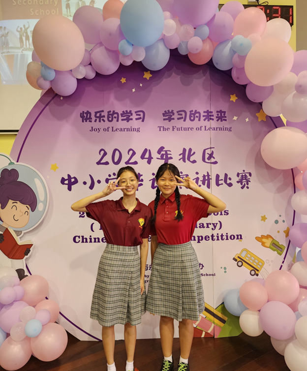 Two girls in red shirts and plaid skirts pose with peace signs at a 2024 Chinese reading competition backdrop with balloons.