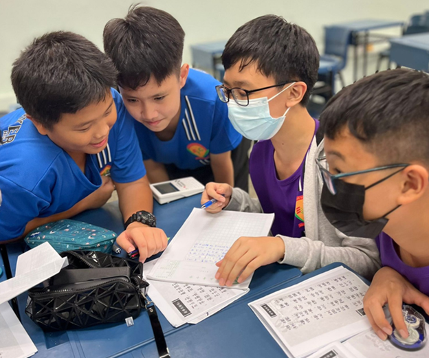 Four young students huddle together over worksheets at a desk, two wearing face masks and glasses.