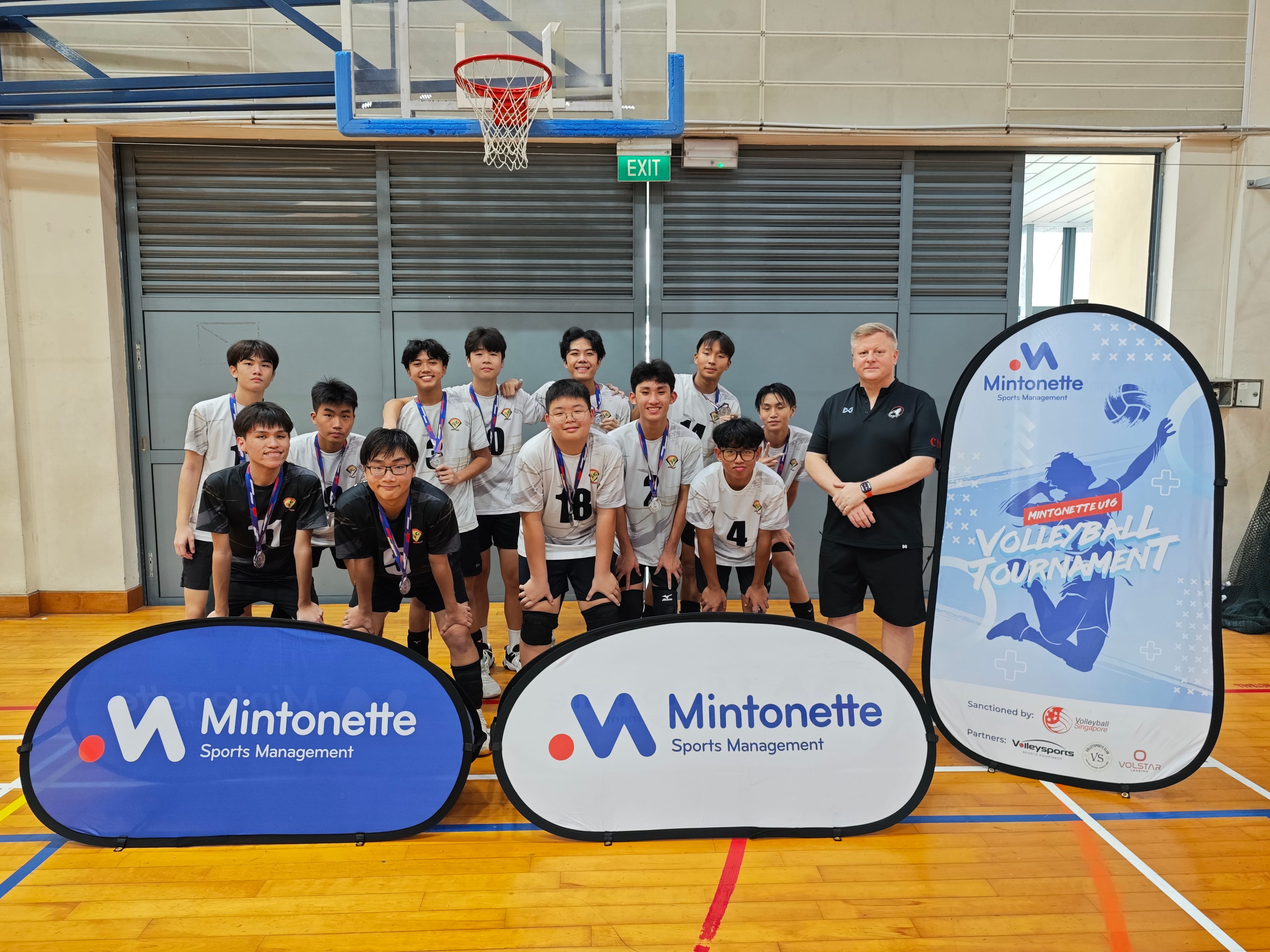 Volleyball team in uniforms pose in a gym behind Mintonnette banners, with a coach beside a tournament sign.