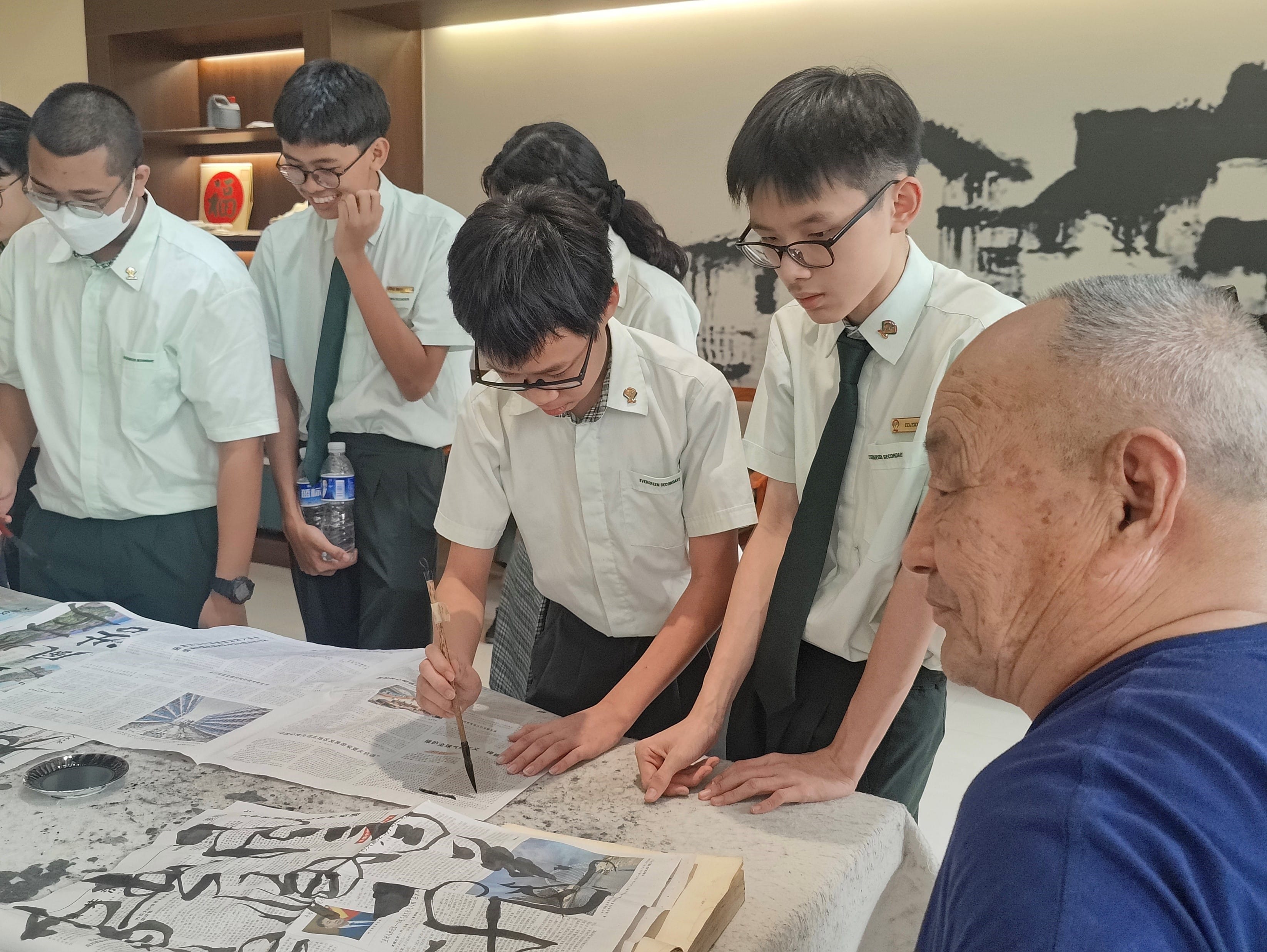 Students in school uniforms watch as one practices Chinese calligraphy with a brush on newspaper, guided by an elderly man.