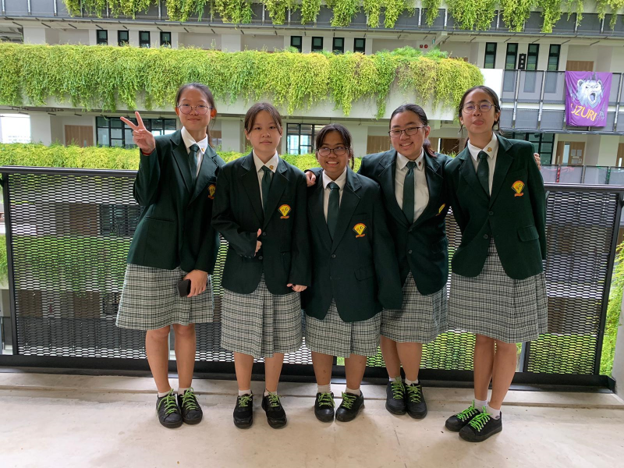 Five girls in dark green school blazers and plaid skirts pose together on a balcony with green ivy-covered walls behind them.