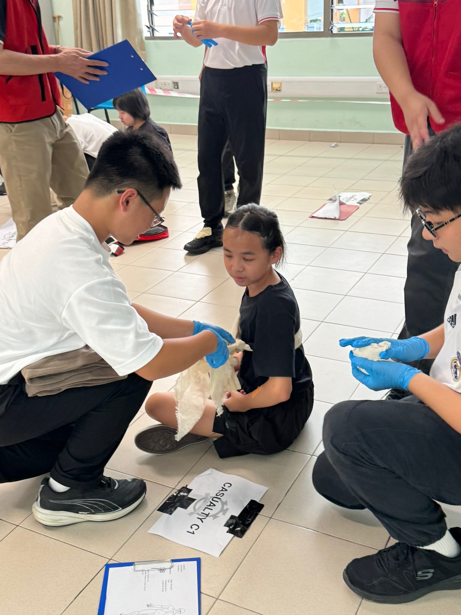 Students in blue gloves practice first aid bandaging on a seated girl during a casualty drill exercise indoors.