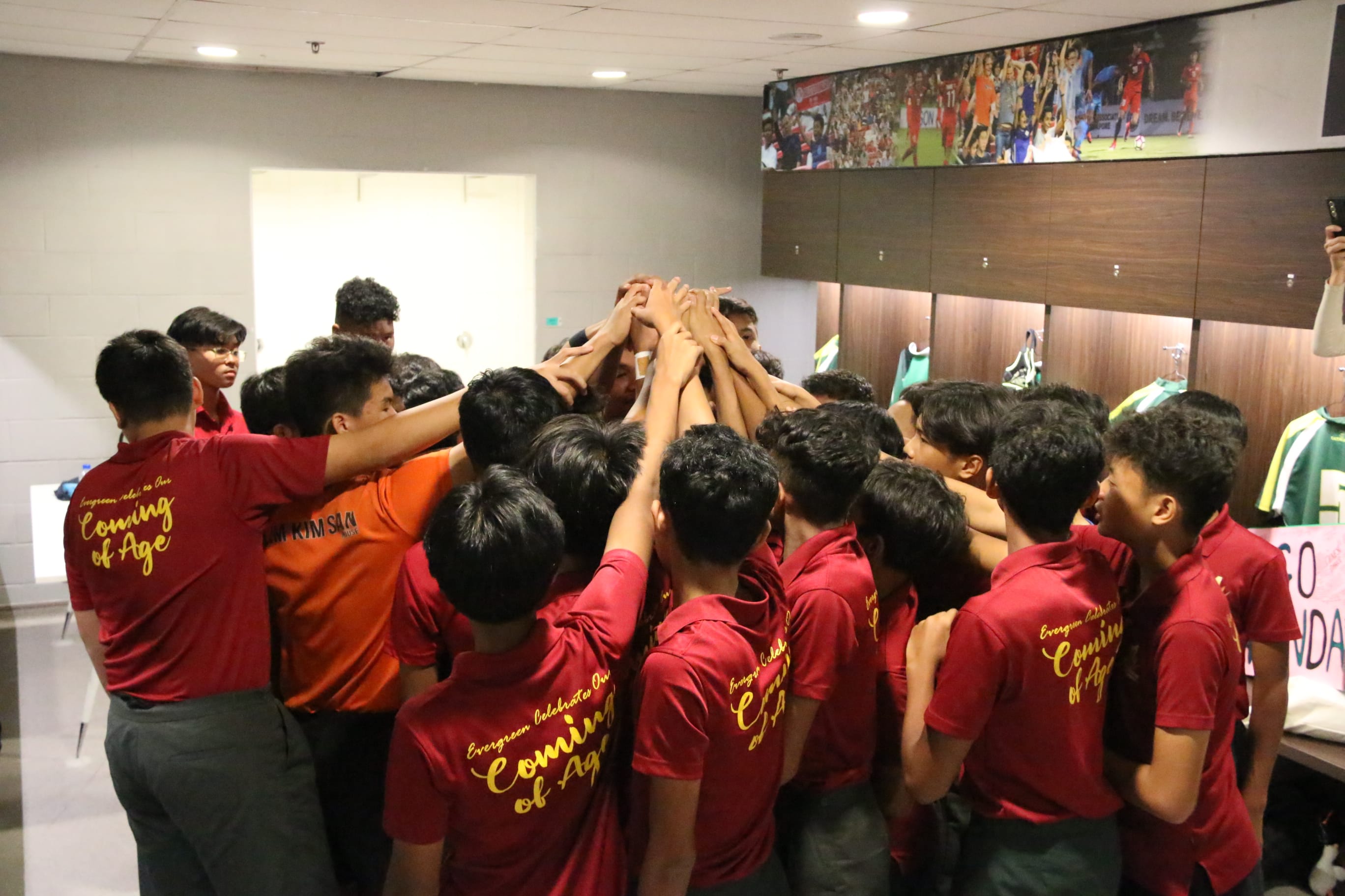 A group of young men in red "Coming of Age" shirts huddle with hands stacked together in a locker room.