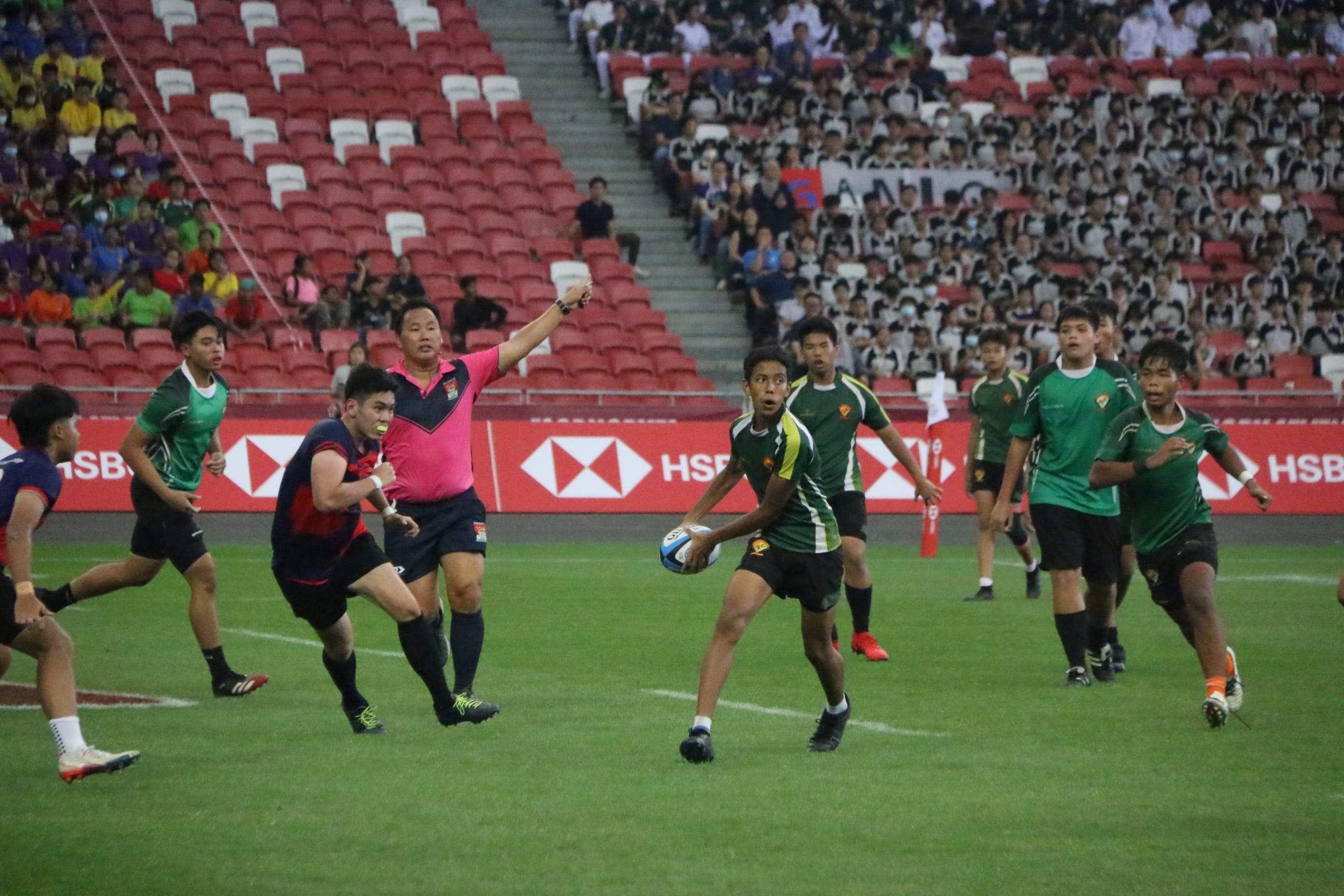 Young rugby players compete on a green field in a stadium with red seats and a large crowd watching.