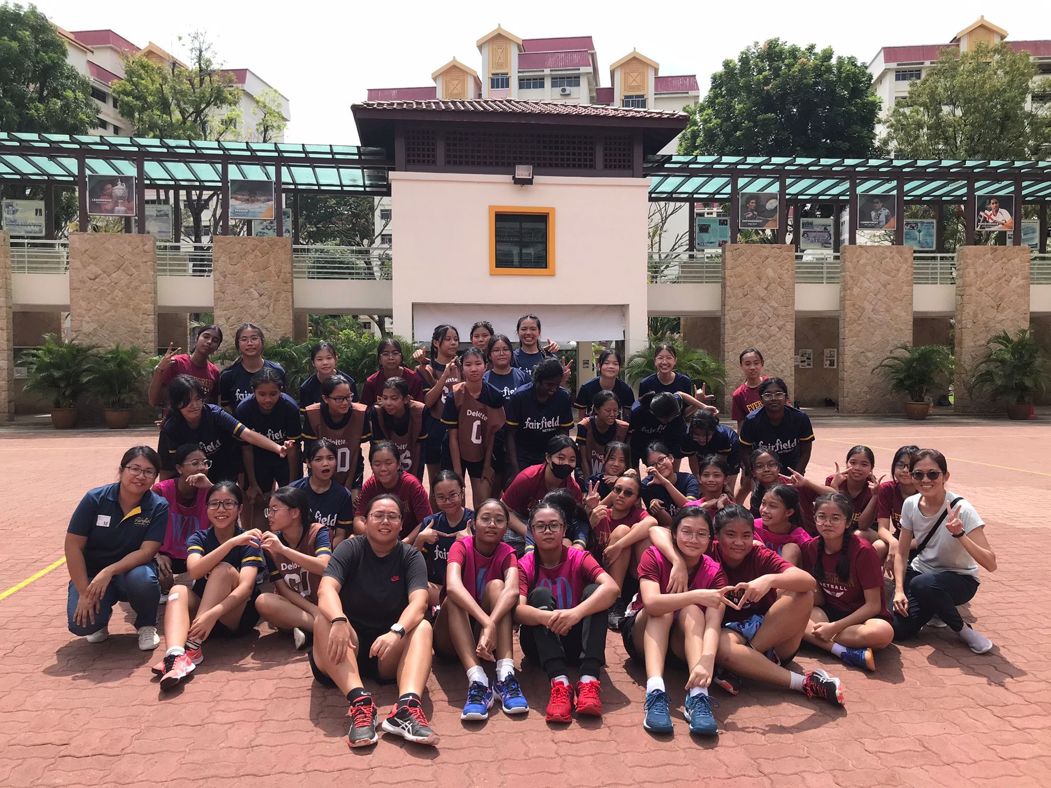 Large group of girls in sports uniforms posing together on an outdoor court in front of a school building.
