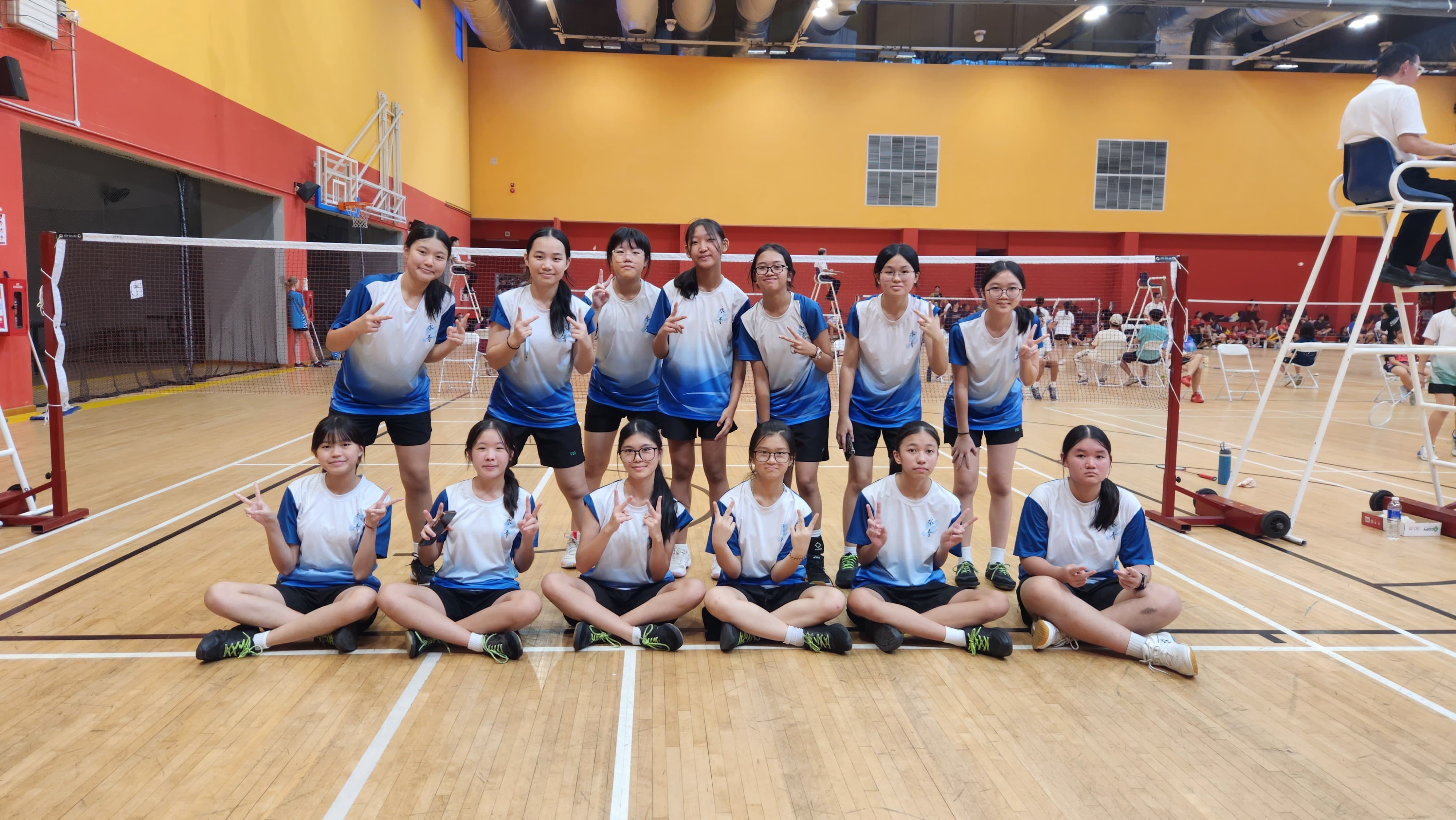 A group of young girls in blue and white jerseys pose with peace signs on a badminton court indoors.