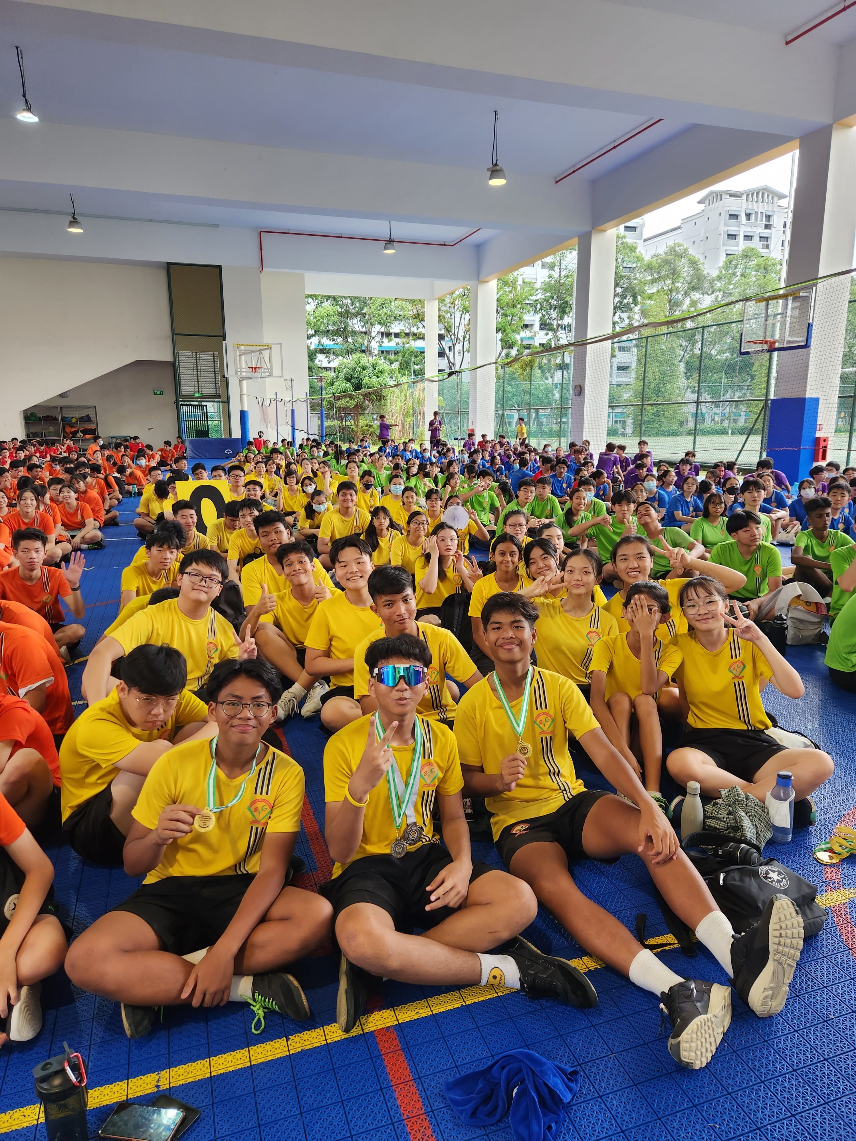 Large group of students in yellow, orange, green, and blue shirts sitting on a sports hall floor, some wearing medals.