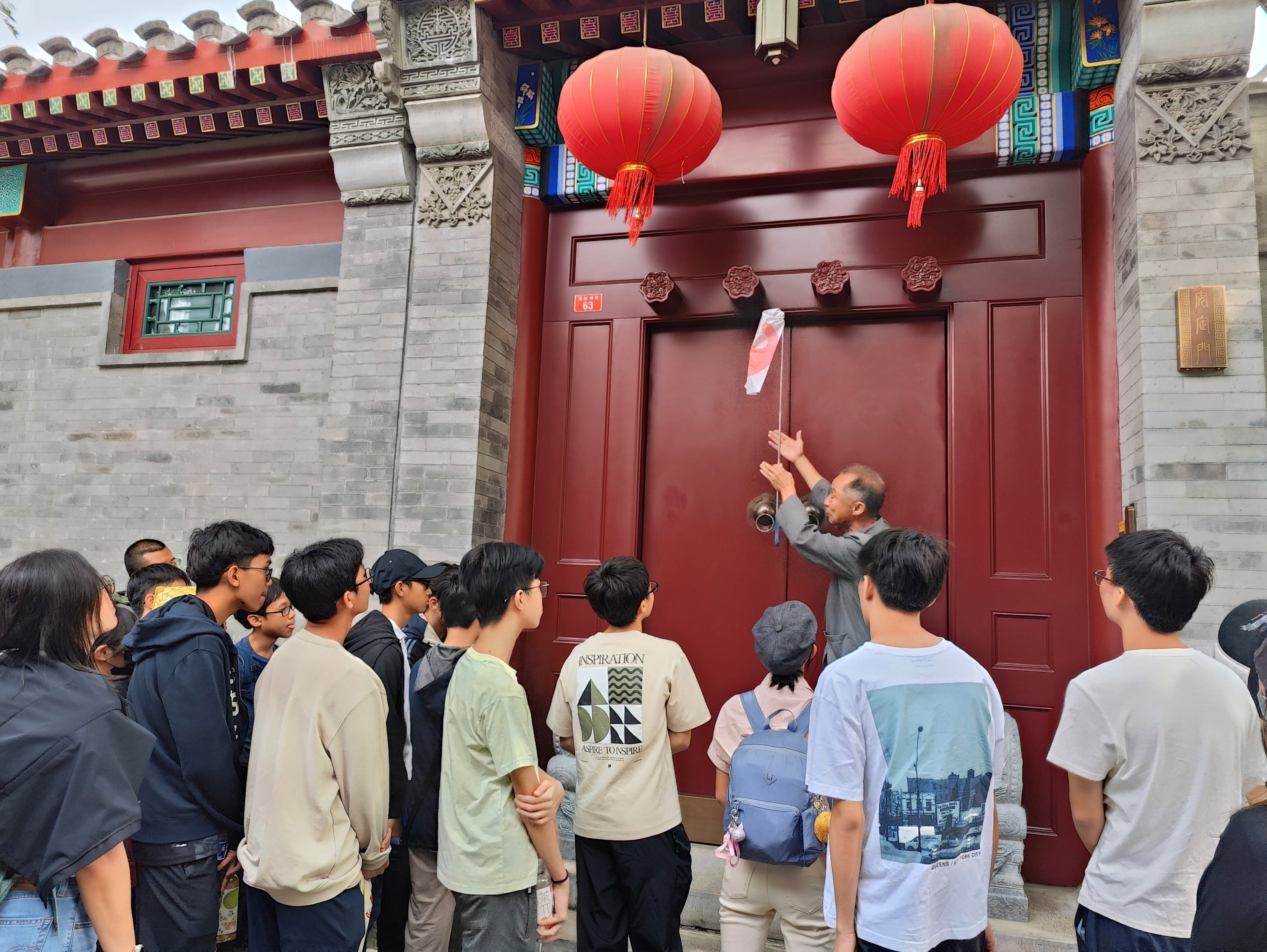 A group watches a man reach up toward a large red Chinese gate decorated with two red hanging lanterns.