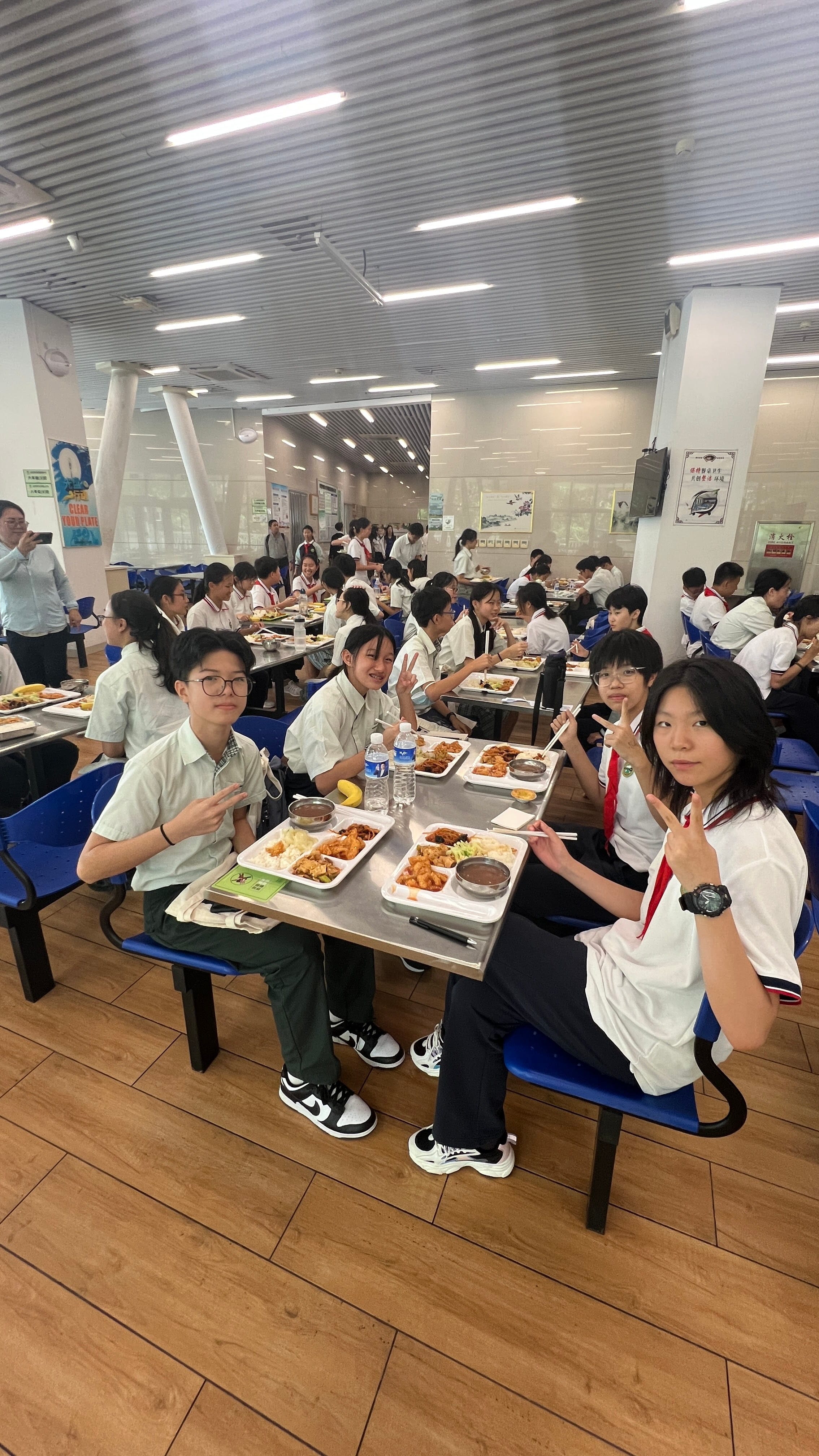 Four students in school uniforms pose with peace signs at a cafeteria table with food trays, surrounded by many other students.
