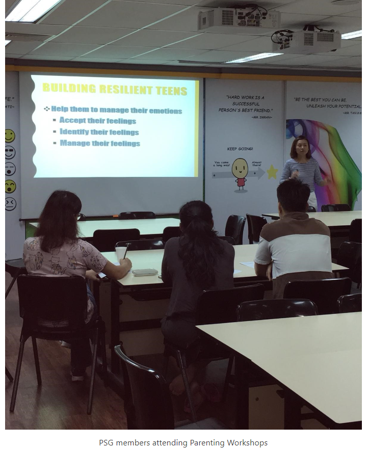 A presenter stands before a projected slide titled "Building Resilient Teens" while three attendees sit at tables facing her.