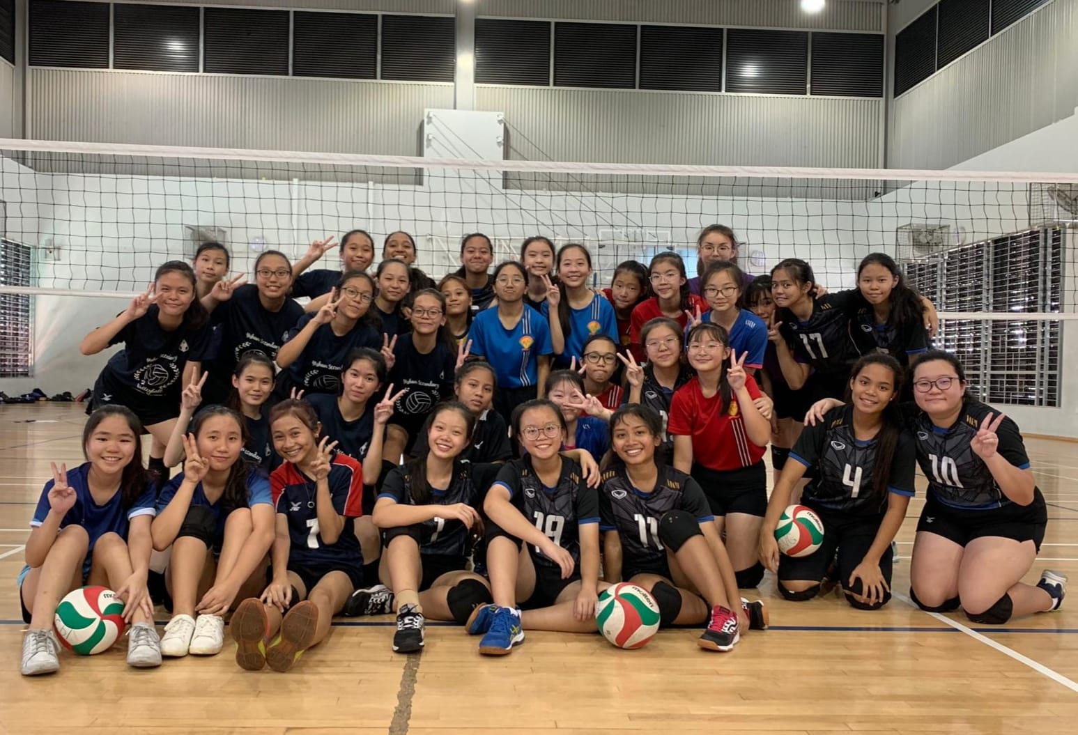 A large group of young female volleyball players pose together in a gym, making peace signs, with volleyballs nearby.