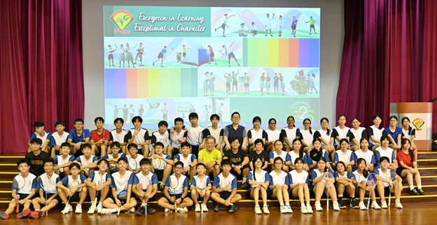 Large group of students and teachers posing on a stage in front of a colorful school presentation screen.
