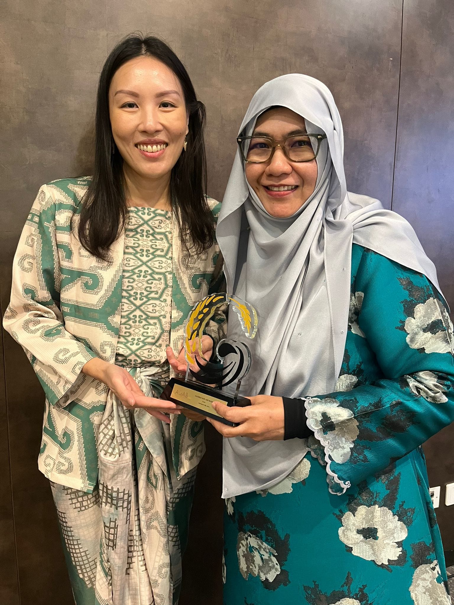Two smiling women holding a decorative glass trophy, one in a patterned jacket, the other in a hijab and teal dress.