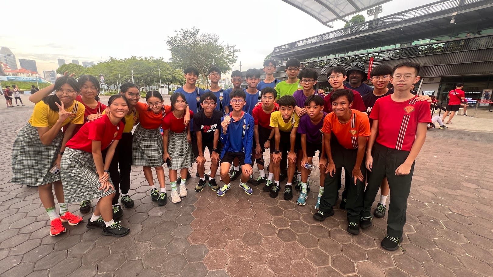 A large group of teenagers in colorful sportswear pose together outdoors on a paved plaza near a stadium.