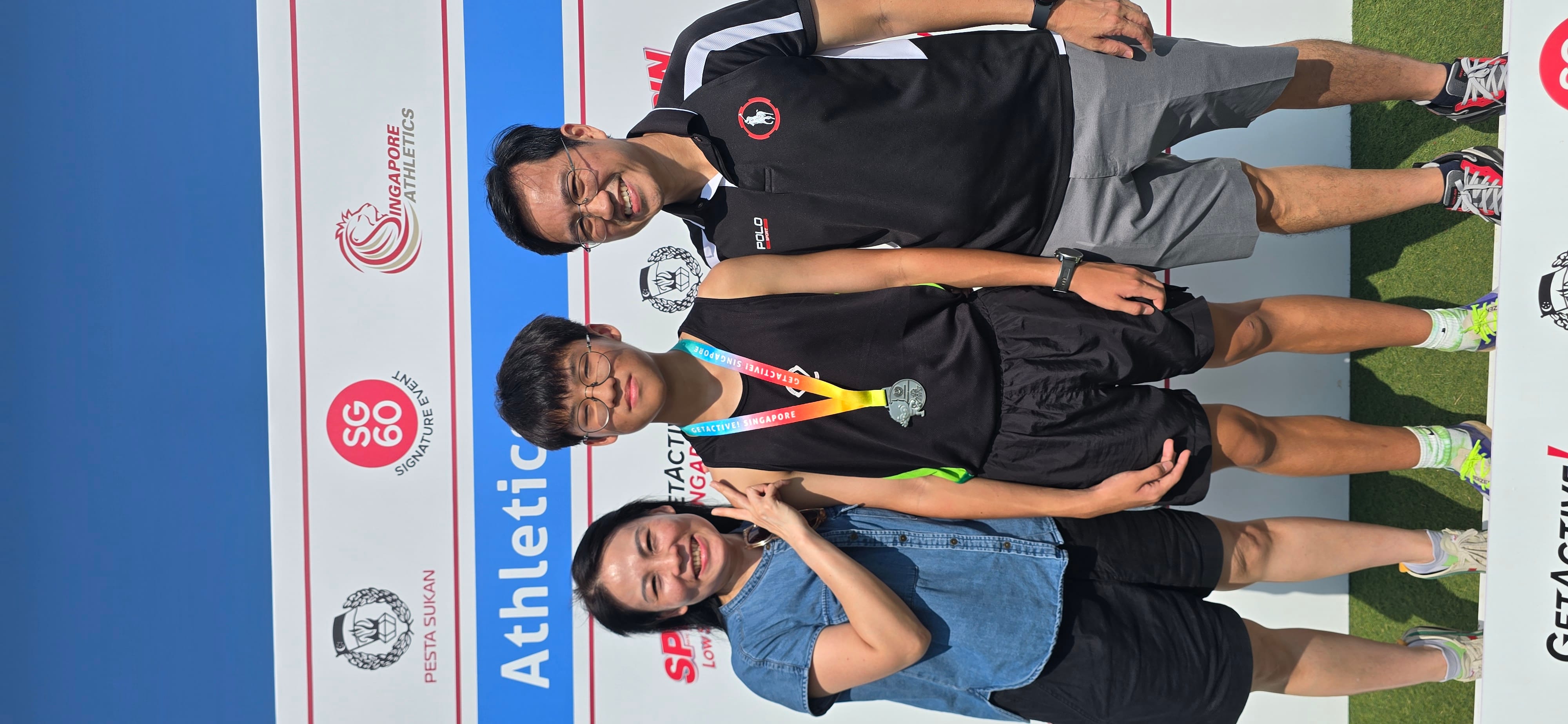 A young athlete wearing a medal stands between two smiling adults at a Singapore Athletics event backdrop.