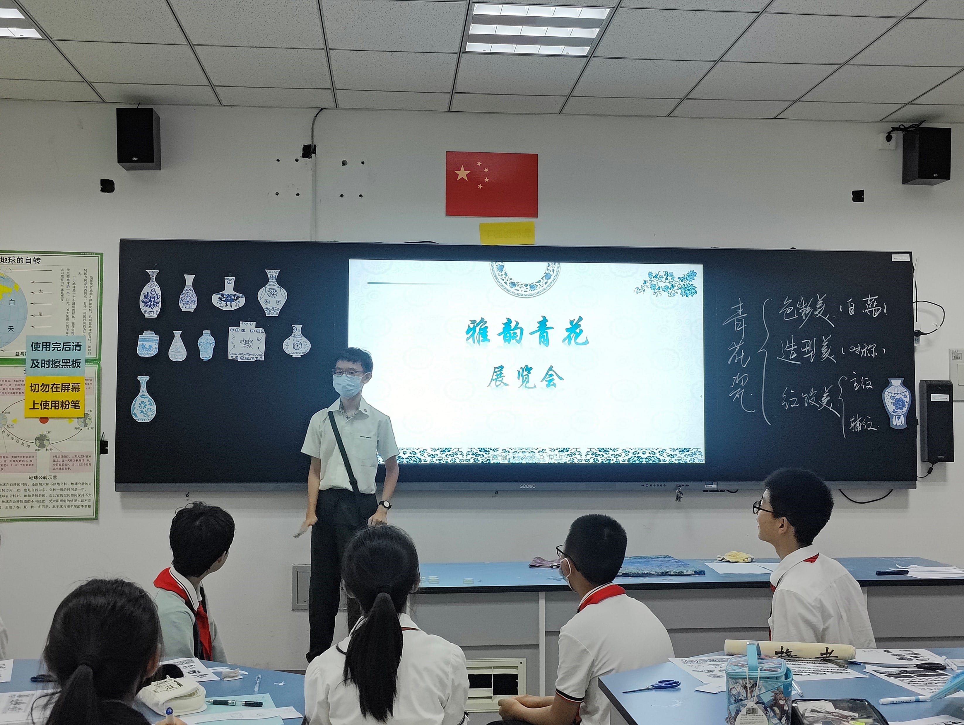 A masked teacher stands at a blackboard displaying a Chinese blue-and-white porcelain exhibition presentation, with students seated facing him.