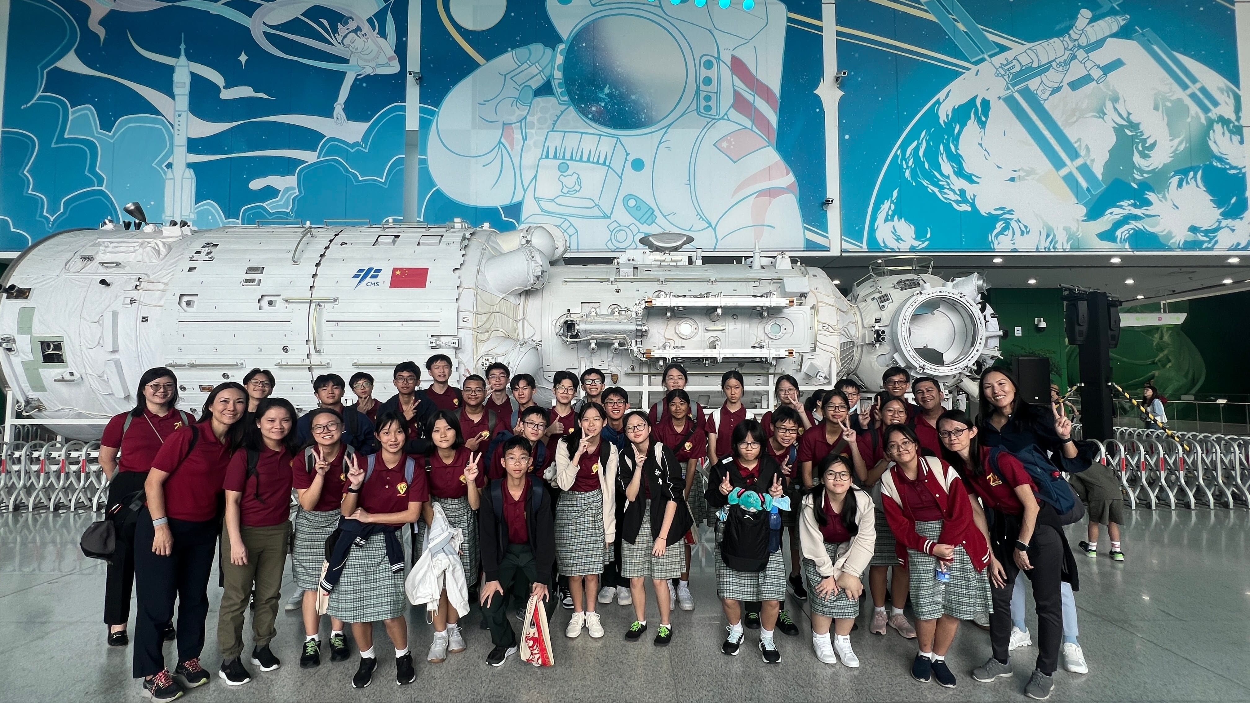 A group of students in red uniforms pose in front of a Chinese space station model and an astronaut mural.