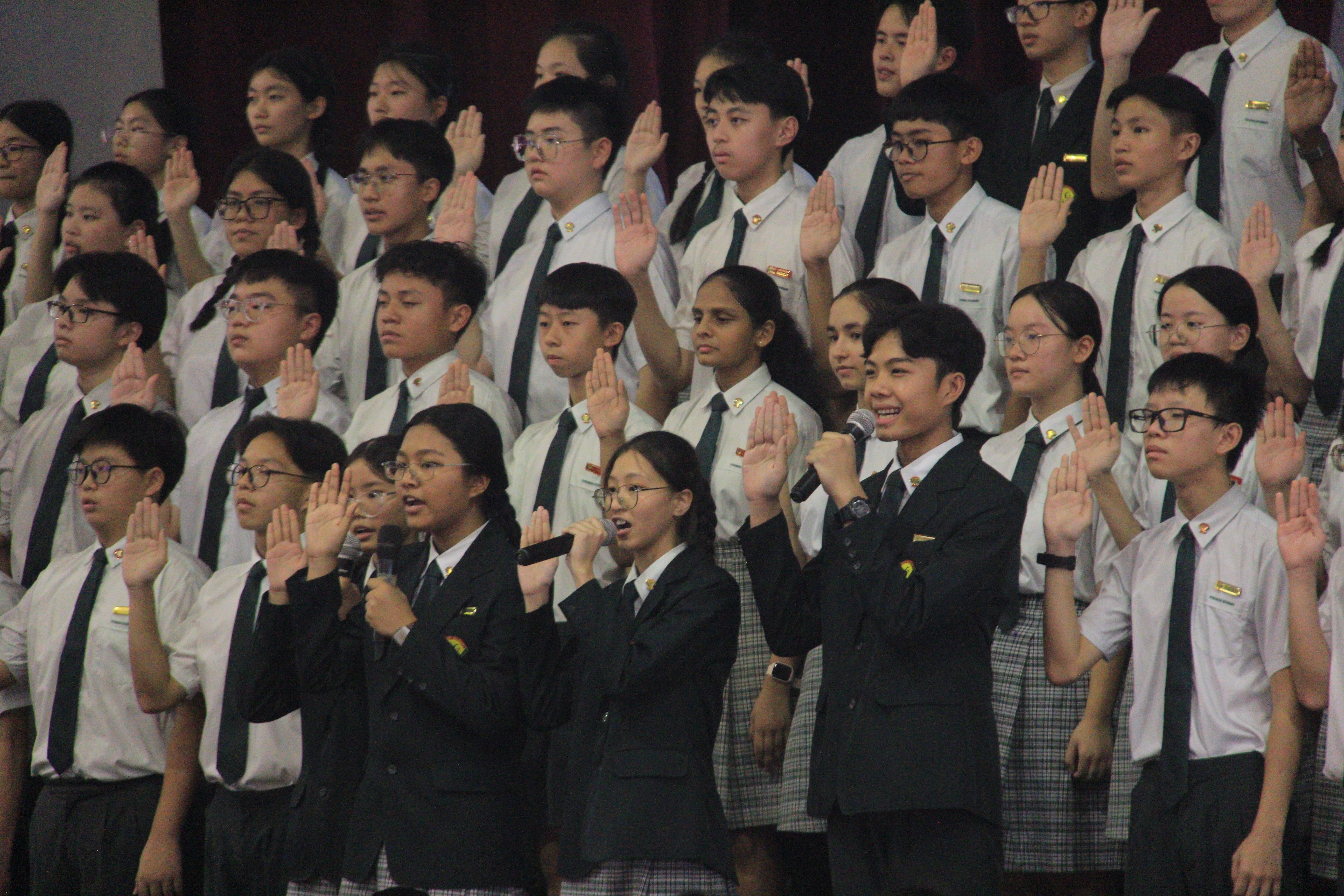 Students in uniforms stand with right hands raised; four in front hold microphones, one smiling.