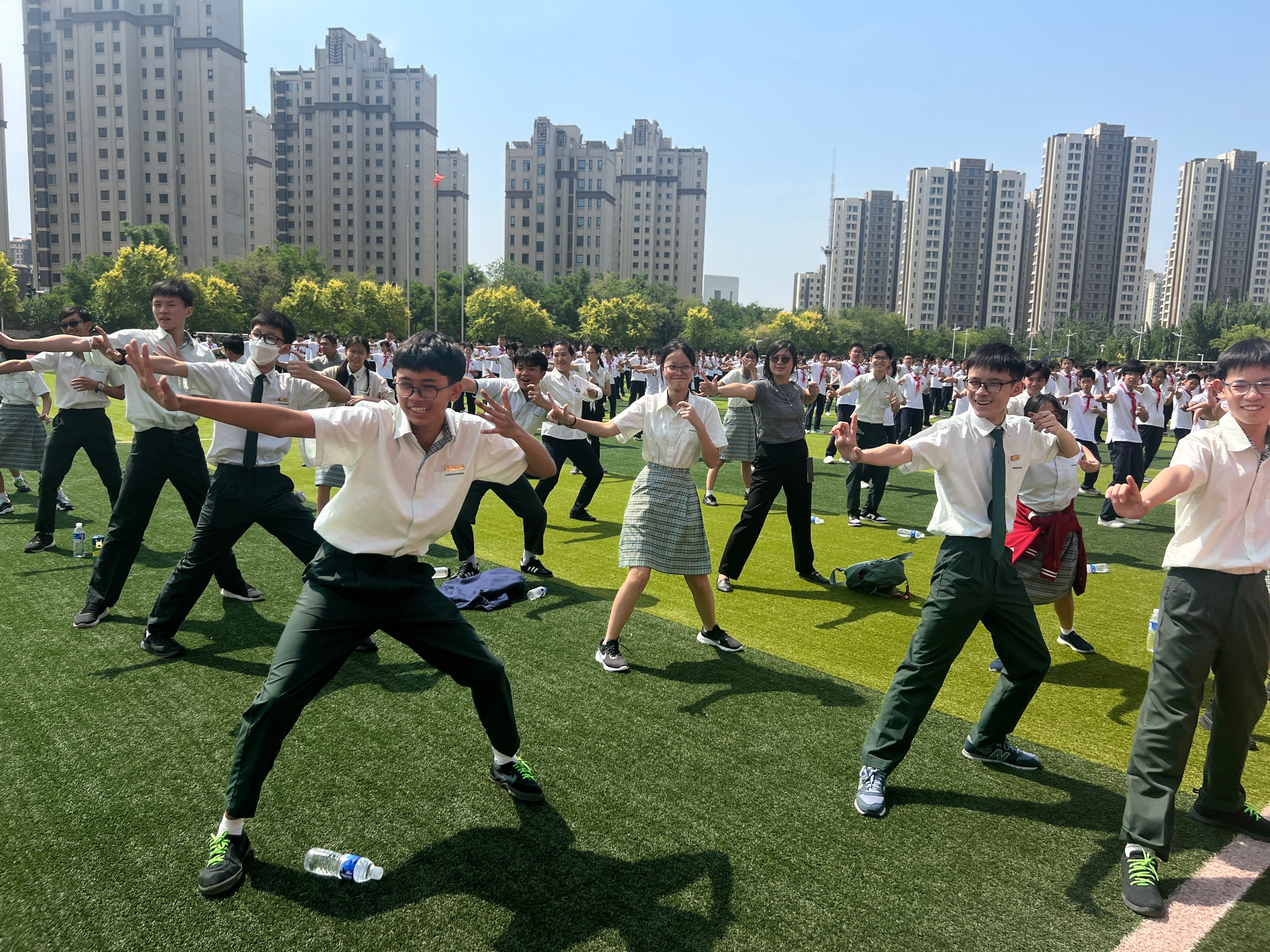 Students in uniforms practice synchronized poses on a sports field, with tall apartments behind.