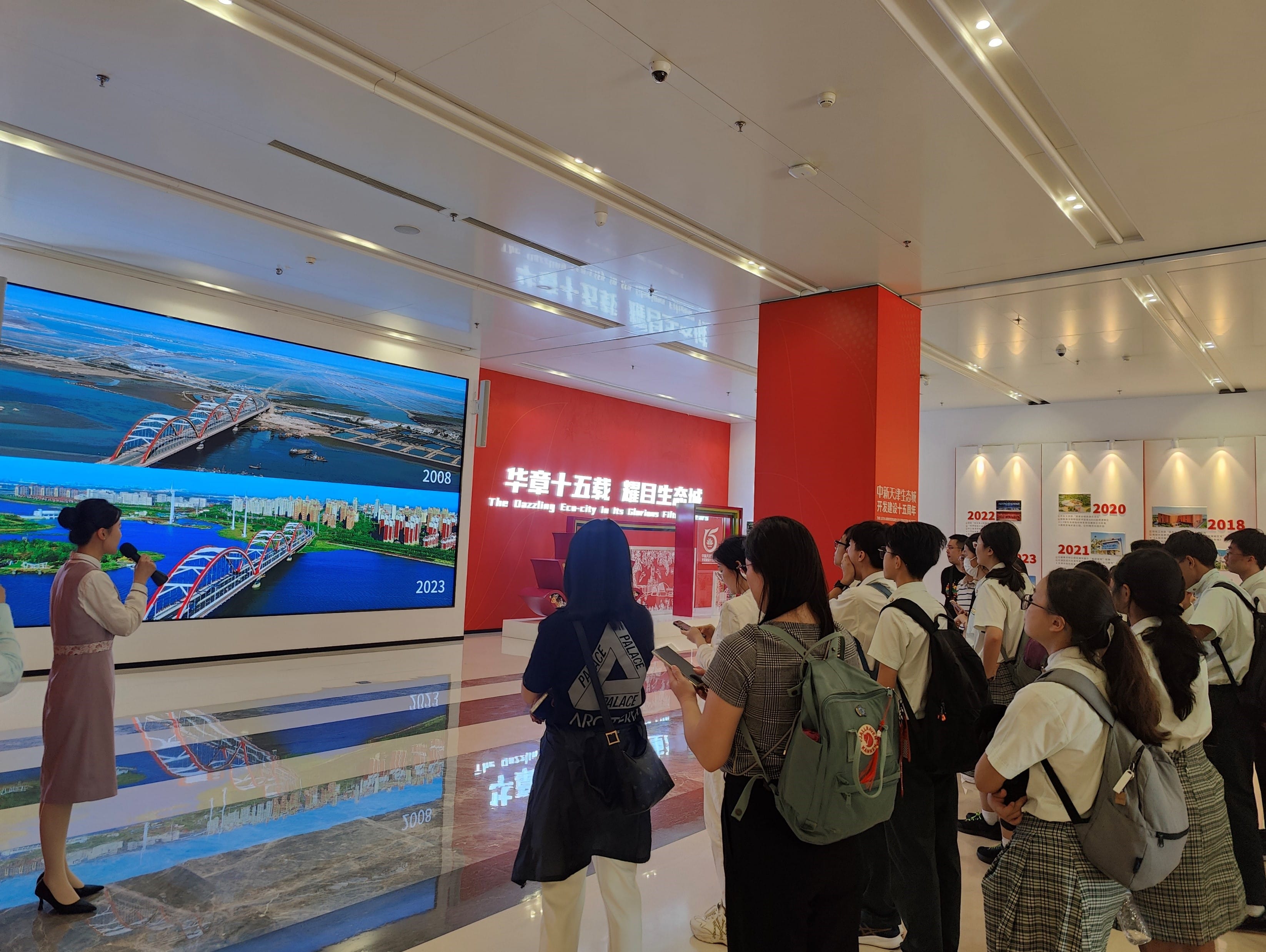 A guide speaks to a group of visitors in an exhibition hall with a large screen showing city aerial views from 2008 and 2023.