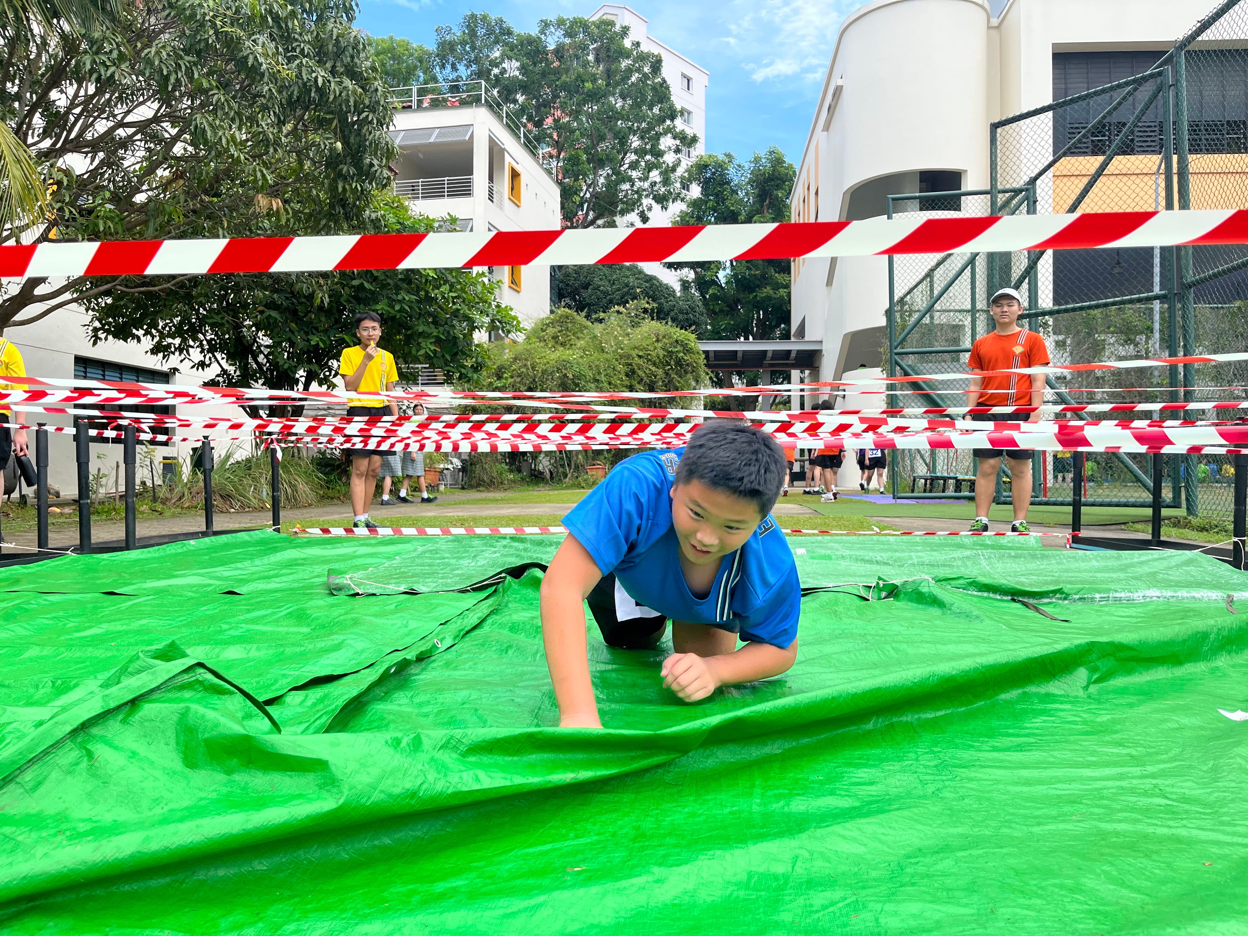 Boy in blue shirt crawling on a green tarp in an outdoor obstacle course area marked with red and white tape.