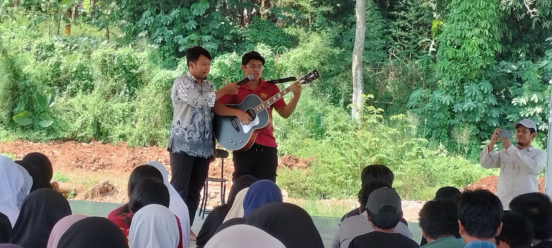 Two men perform outdoors, one playing acoustic guitar and singing into a mic, before a seated audience with trees behind.