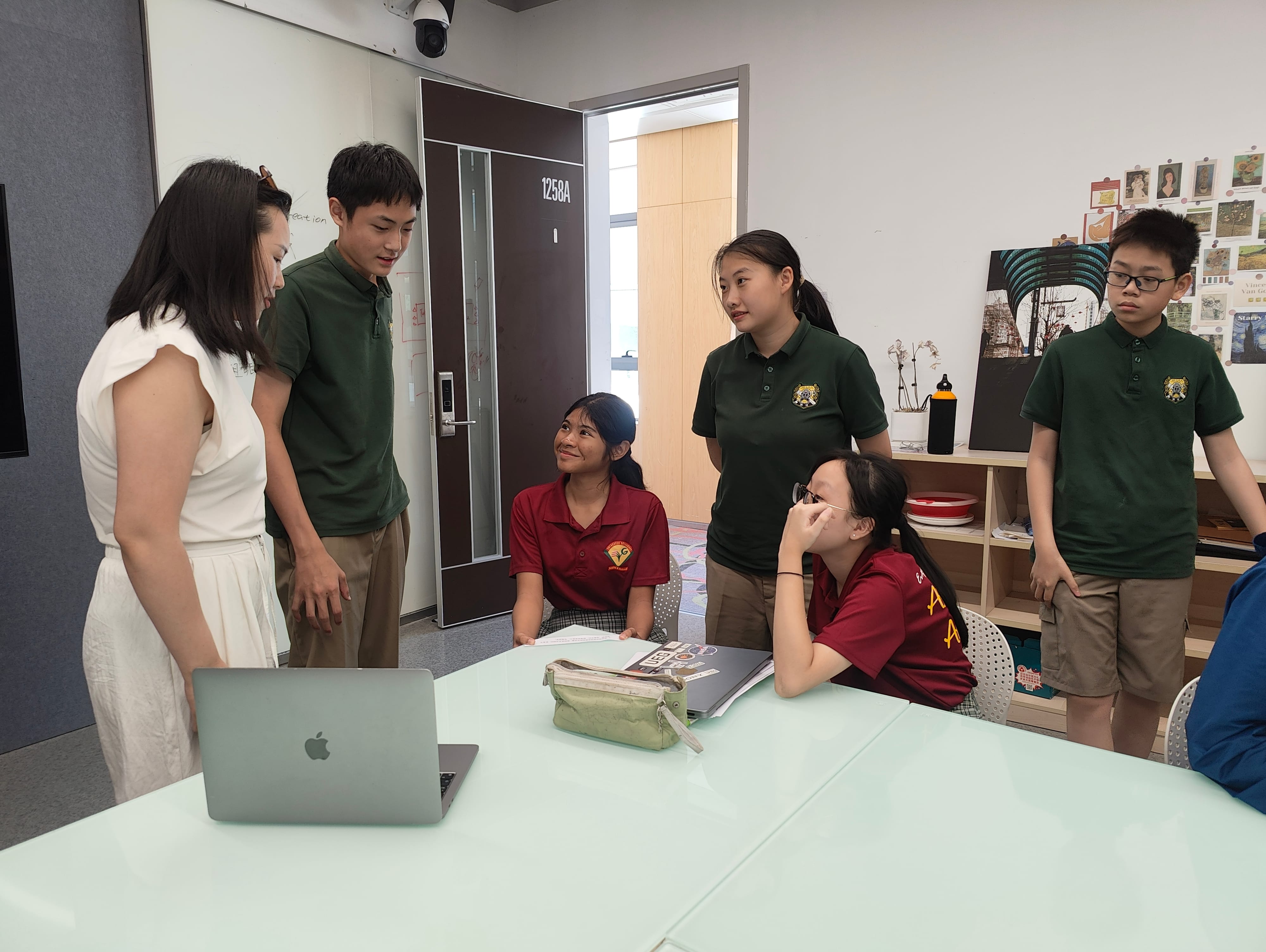 Teacher in white talks with students in green and red uniforms around a table with a laptop.