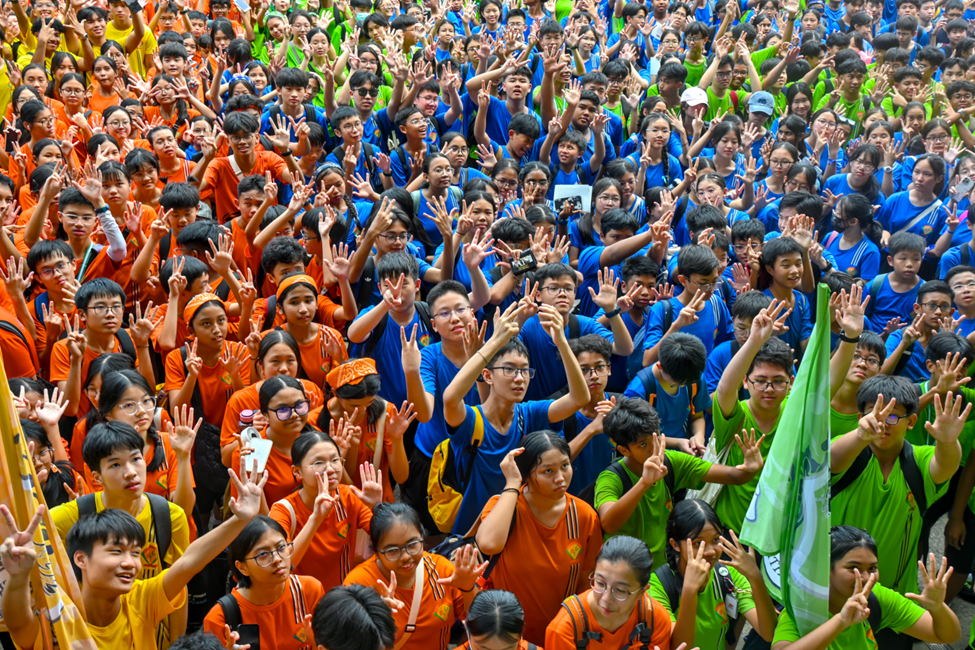 Large crowd of students in orange, blue, green, and yellow shirts waving and raising hands together outdoors.