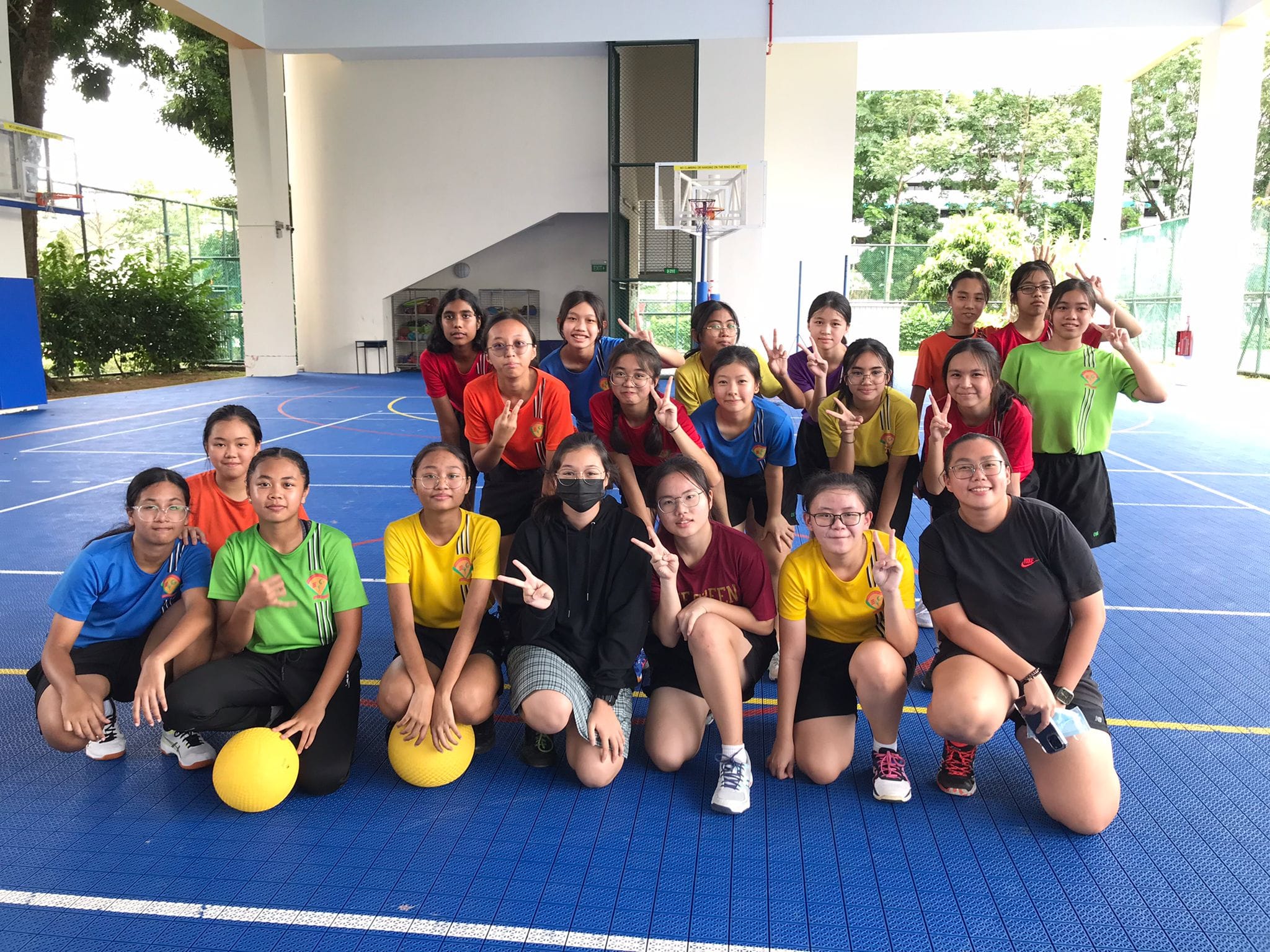 A group of young female students posing on a blue indoor basketball court, making peace signs, with yellow balls nearby.