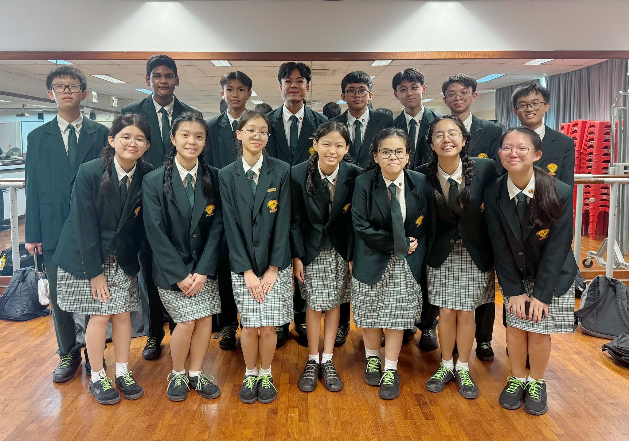 Group of school students in dark green blazers and plaid skirts or dark trousers posing together indoors.