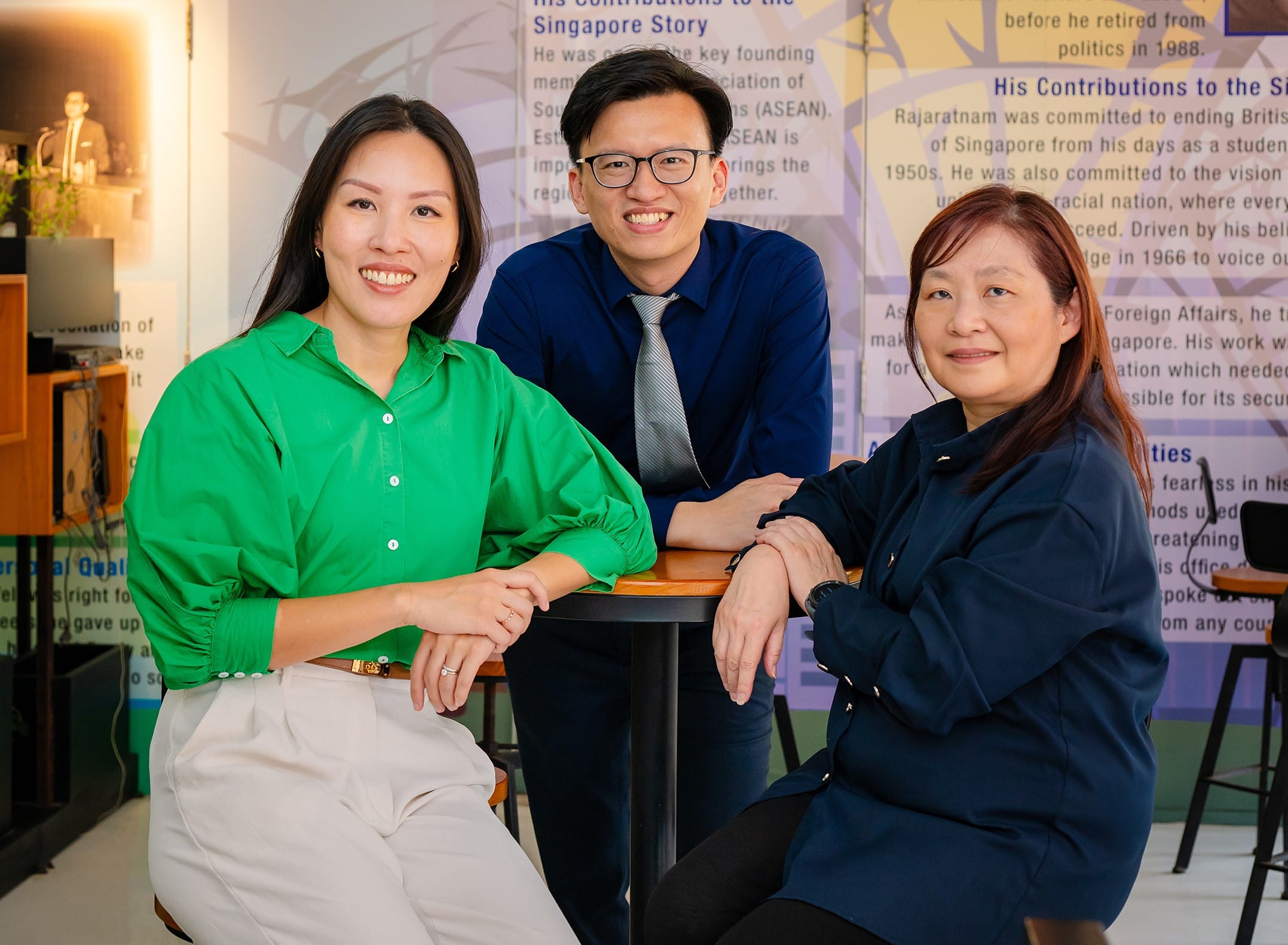 Three smiling people pose at a table in front of a wall display about the Singapore Story.