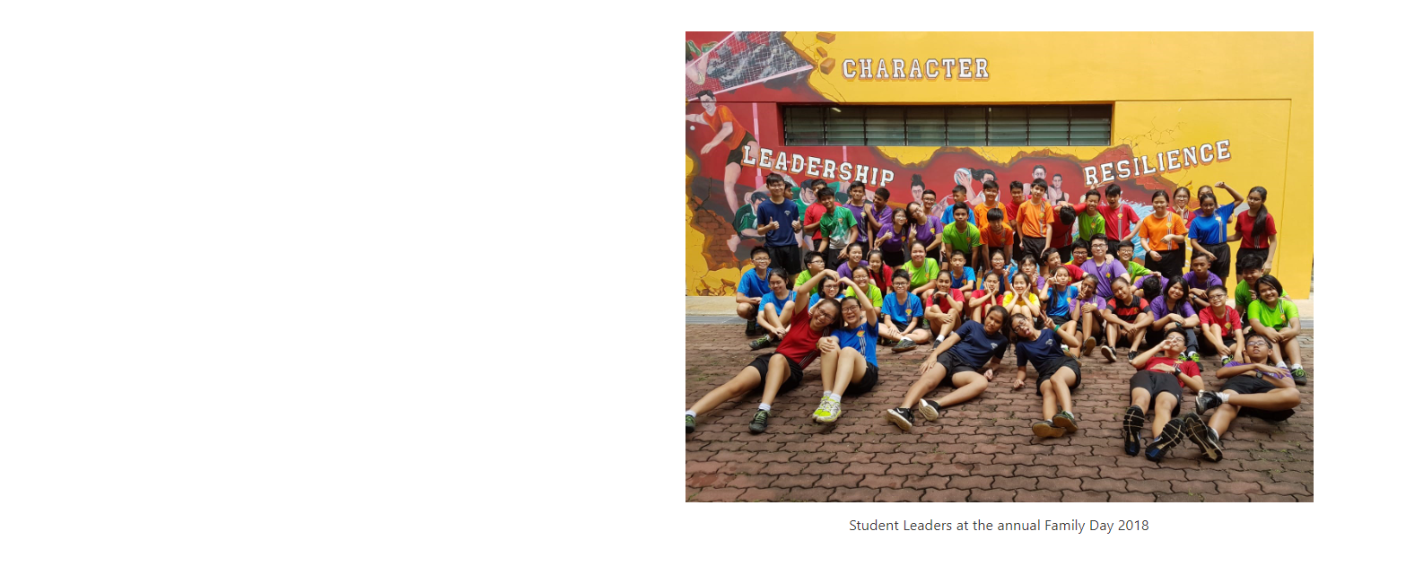 Large group of students posing in front of a yellow wall mural reading Character, Leadership, Resilience.