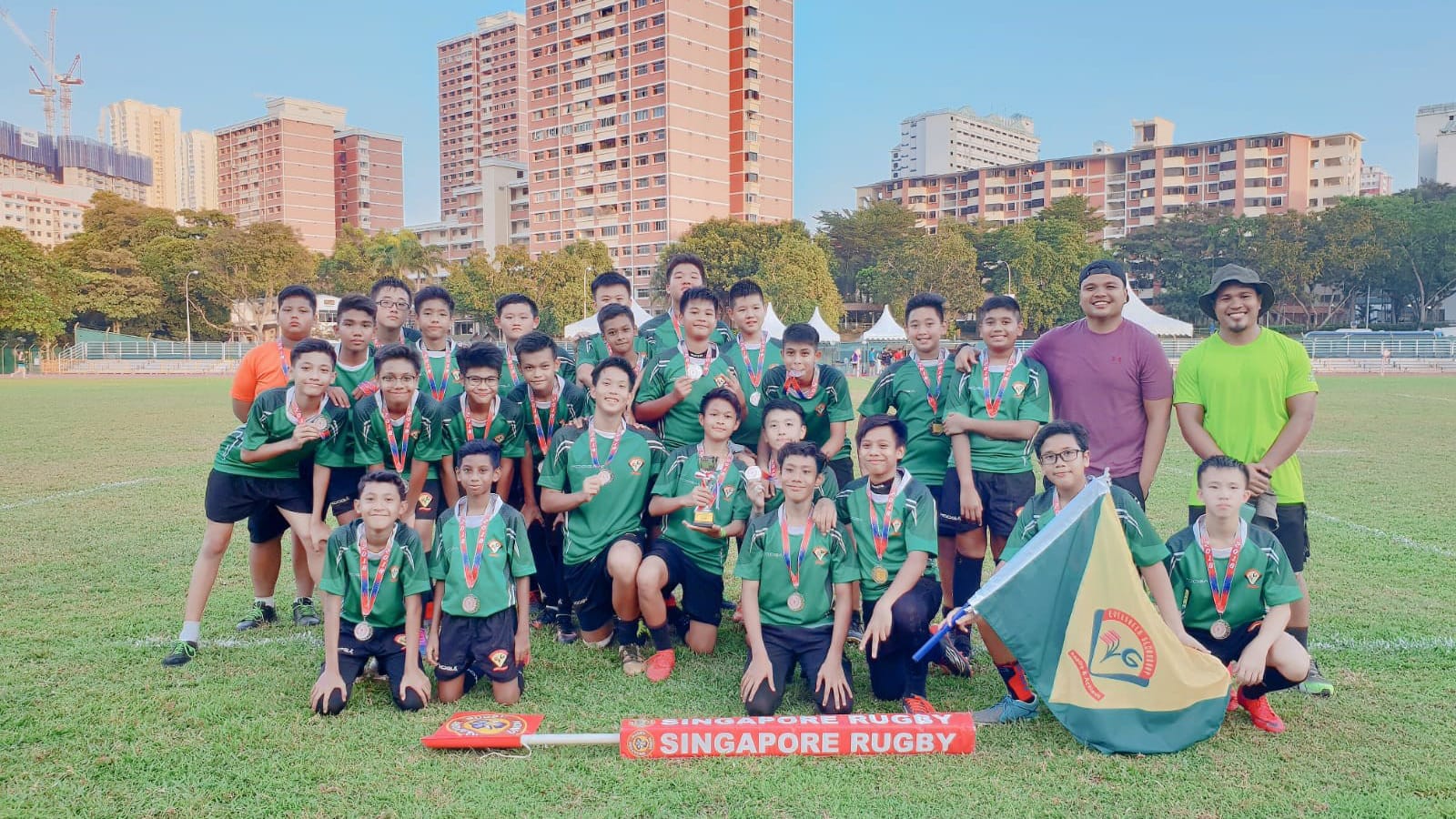 Youth rugby team in green jerseys posing with medals, a trophy, and a Singapore Rugby banner on a field.