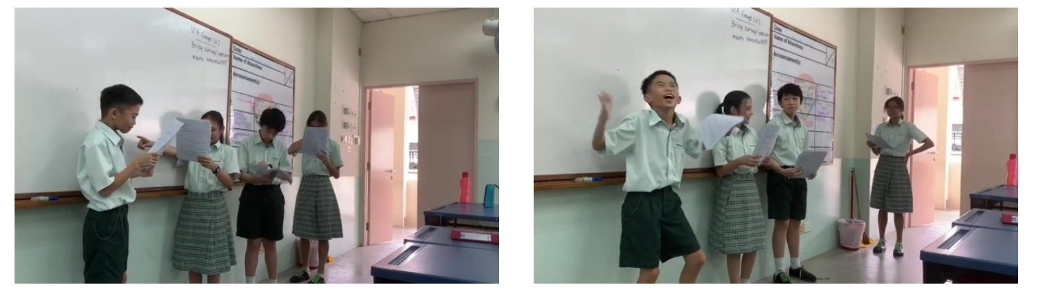 Students in green uniforms stand before a whiteboard, reading papers; one boy gestures expressively with arm raised.