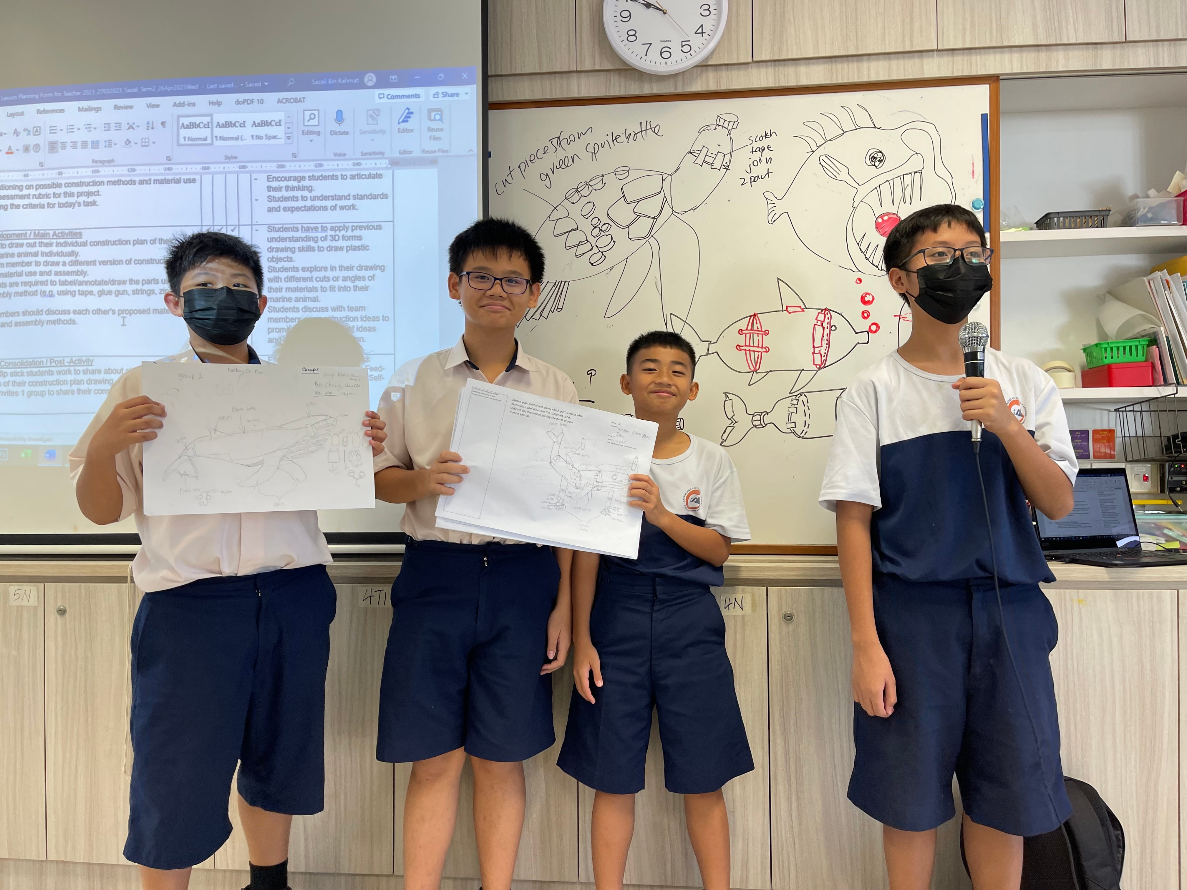 Four school boys in uniforms present drawings of marine animals in a classroom, one holding a microphone.