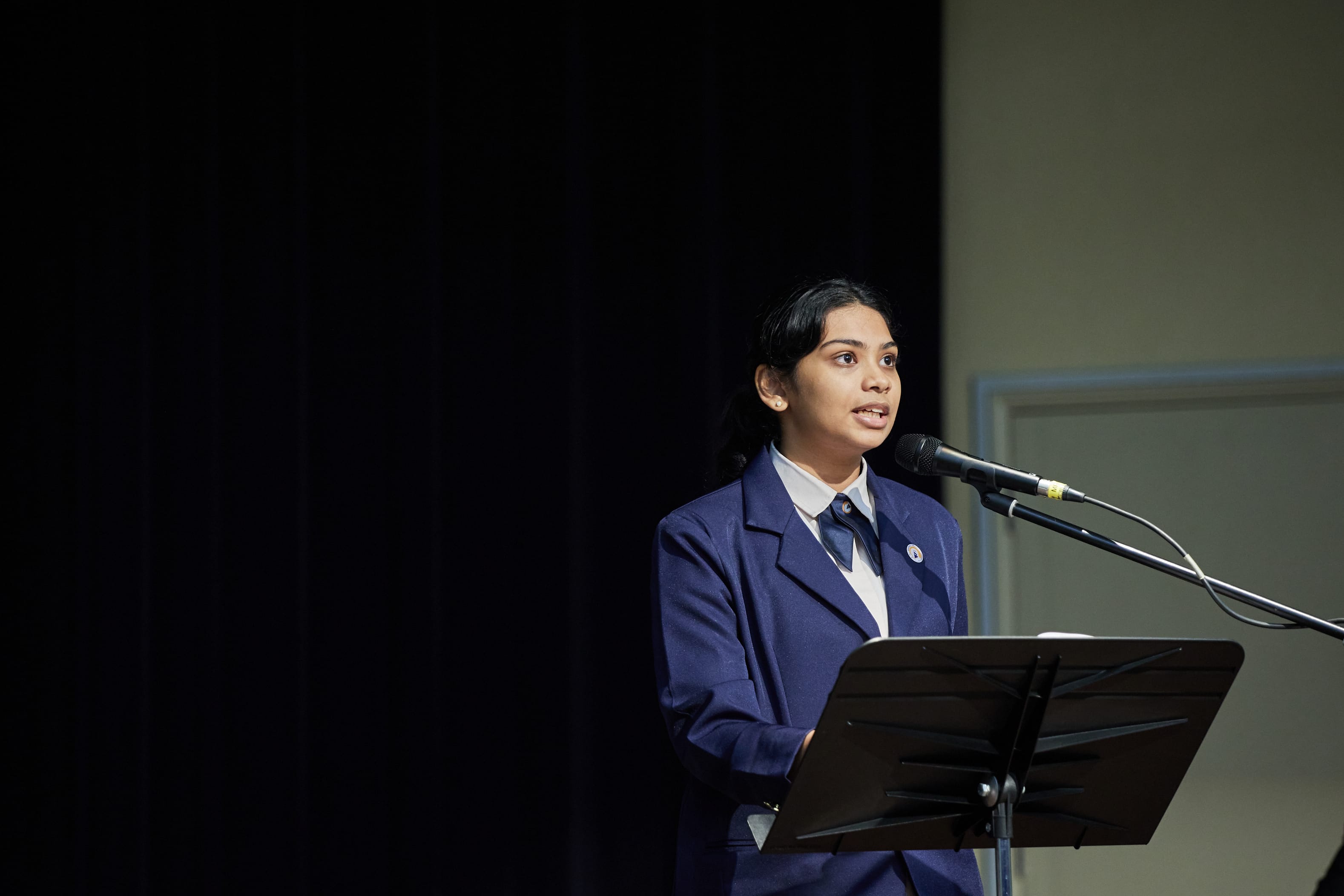 Student in navy blazer speaks into a microphone at a lectern on a stage with dark curtain backdrop.