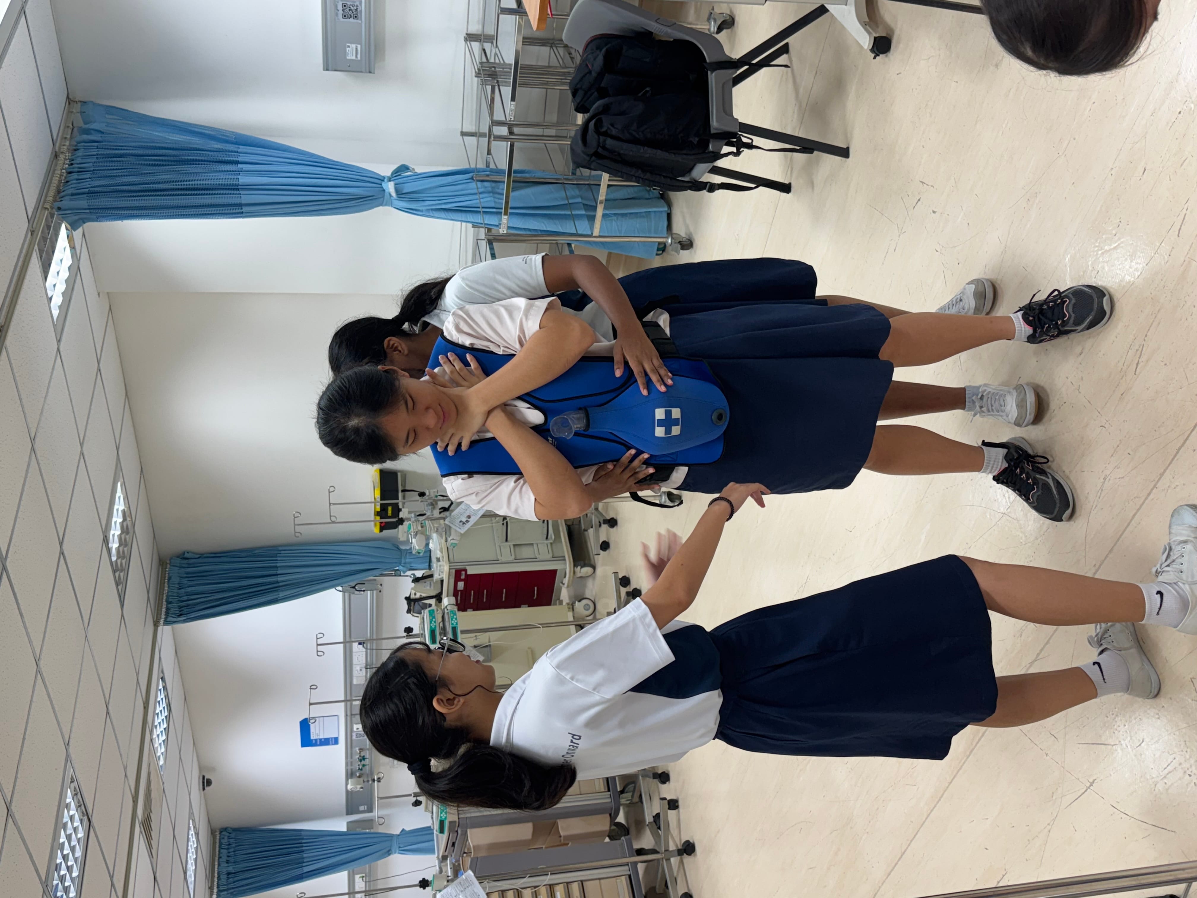 Three school girls in uniforms practice first aid in a medical training room, one wearing a blue CPR vest.