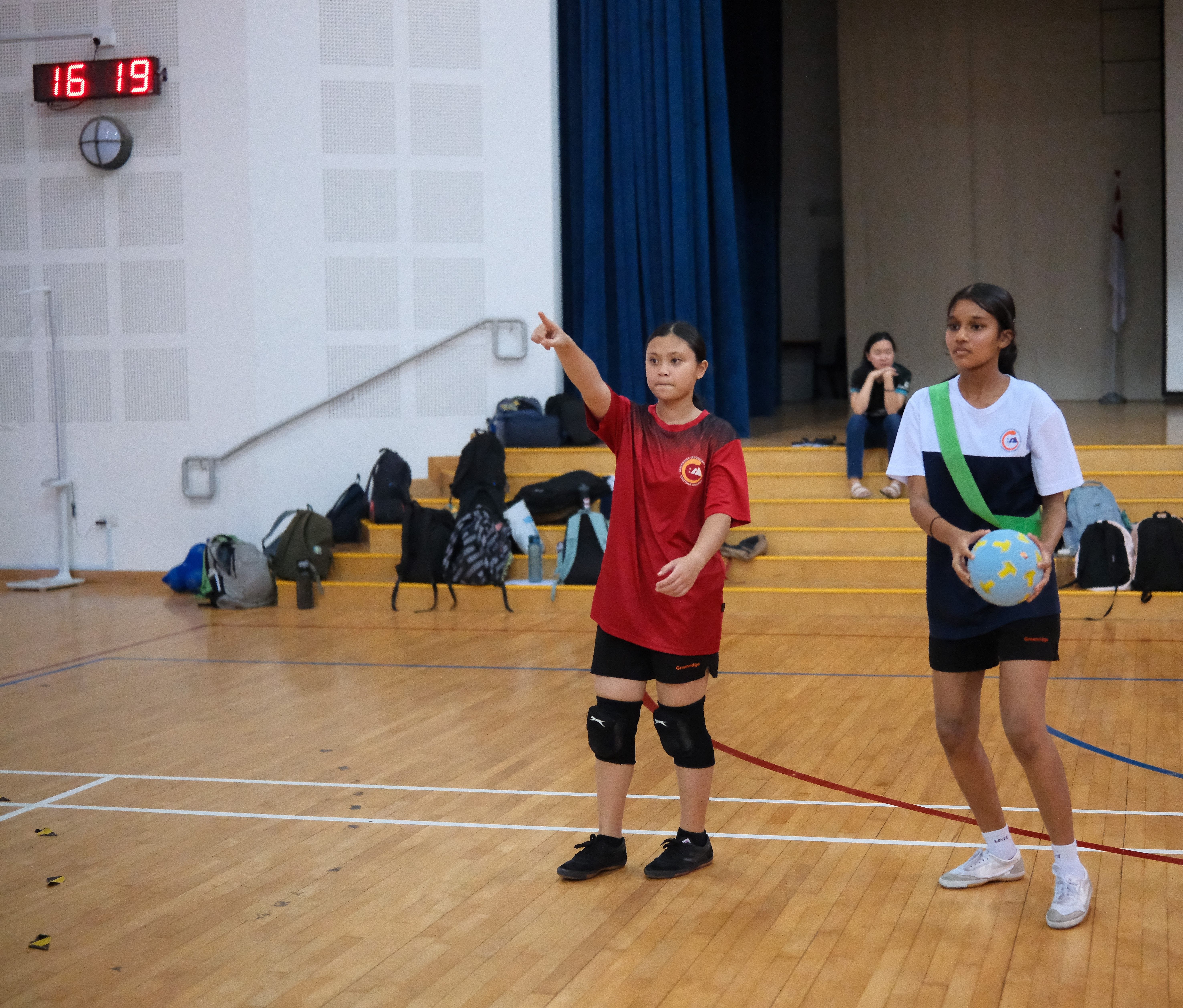 Two girls on an indoor court; one points ahead while the other holds a ball; scoreboard reads 16 19.