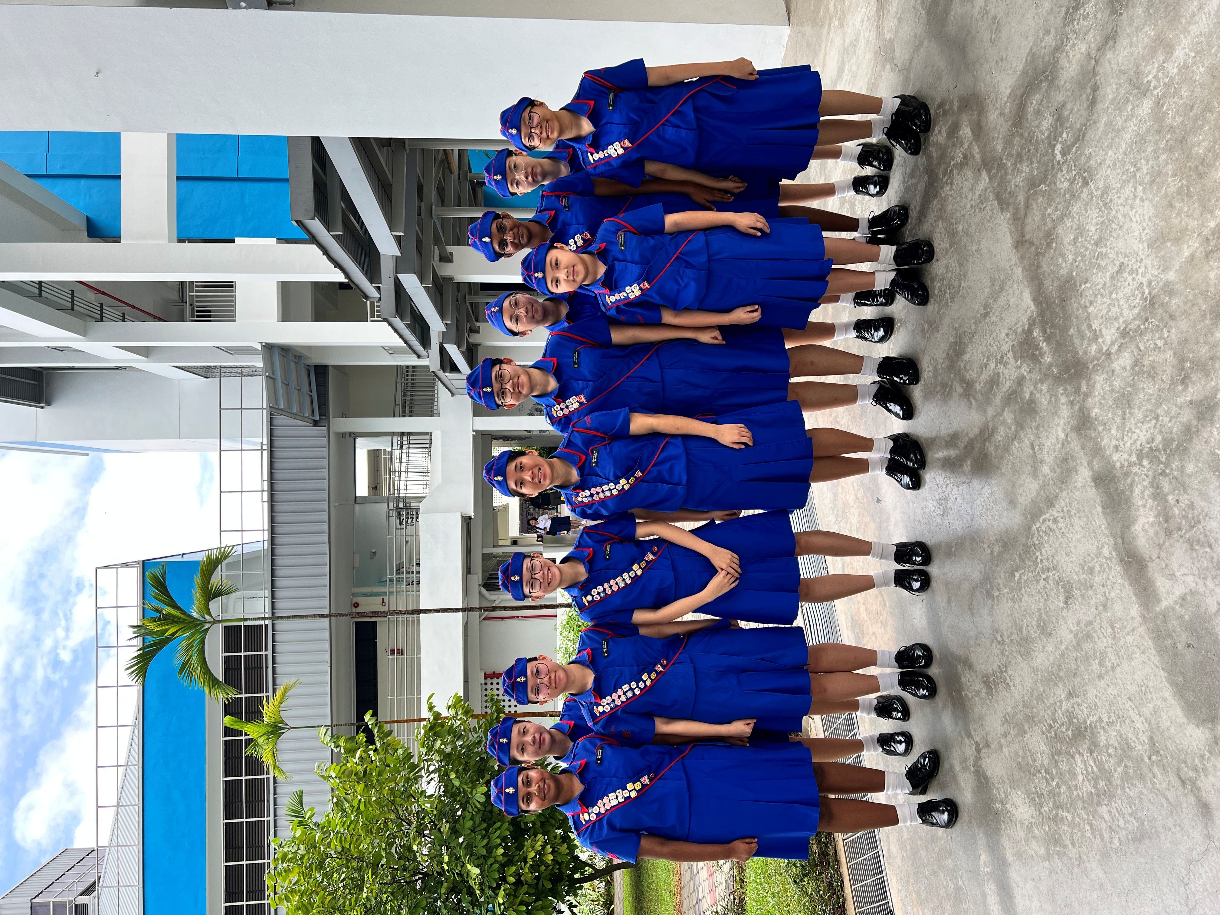 Eleven girls in blue uniforms with caps and badge sashes stand together outdoors near a school building.