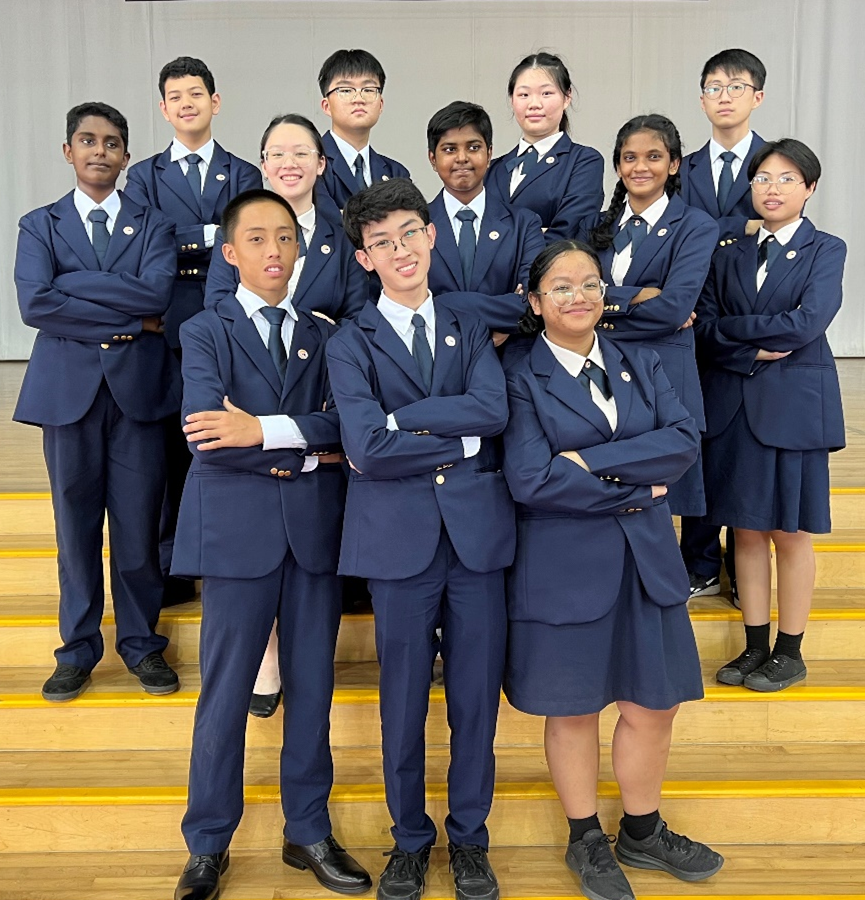 Twelve students in matching navy blue blazers and ties pose with arms crossed on wooden bleachers.