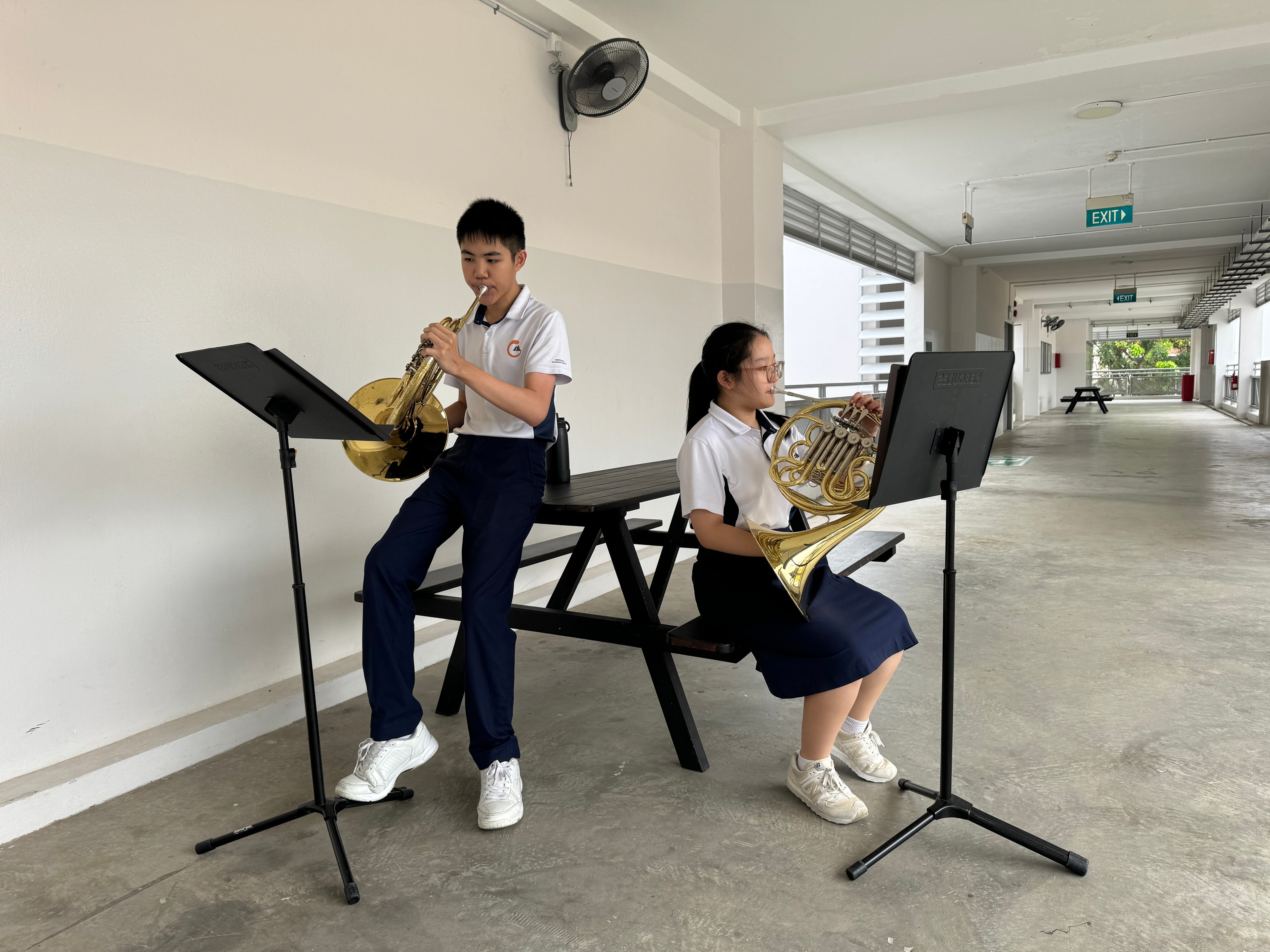Two students in school uniforms practice French horn instruments at music stands in an outdoor school corridor.