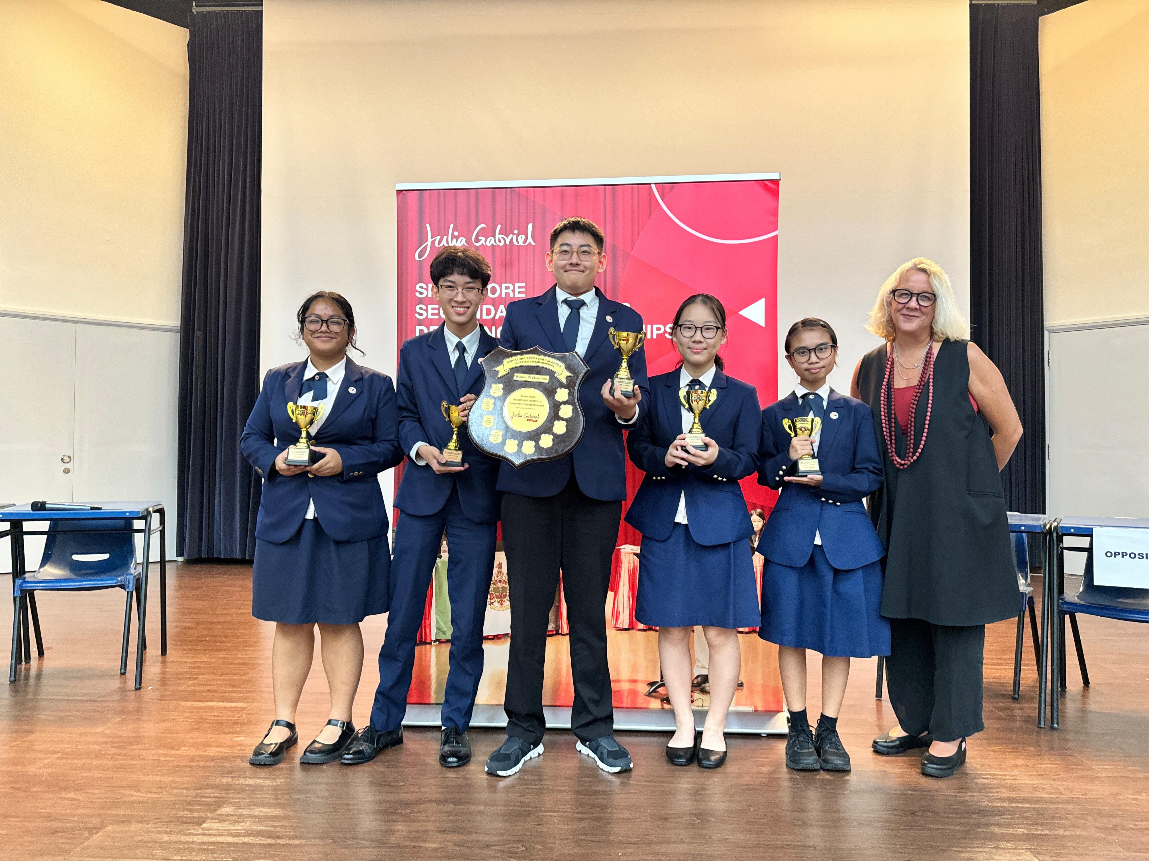 Four students in navy uniforms hold trophies and a shield plaque on stage at a Julia Gabriel debating championship event.