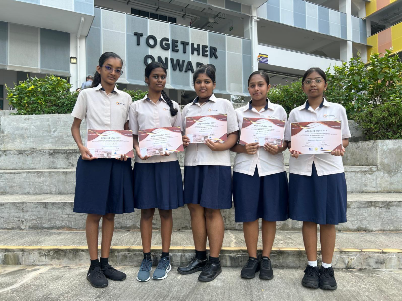 Five schoolgirls in uniforms stand on steps holding certificates, with a "Together Onwards" sign behind them.