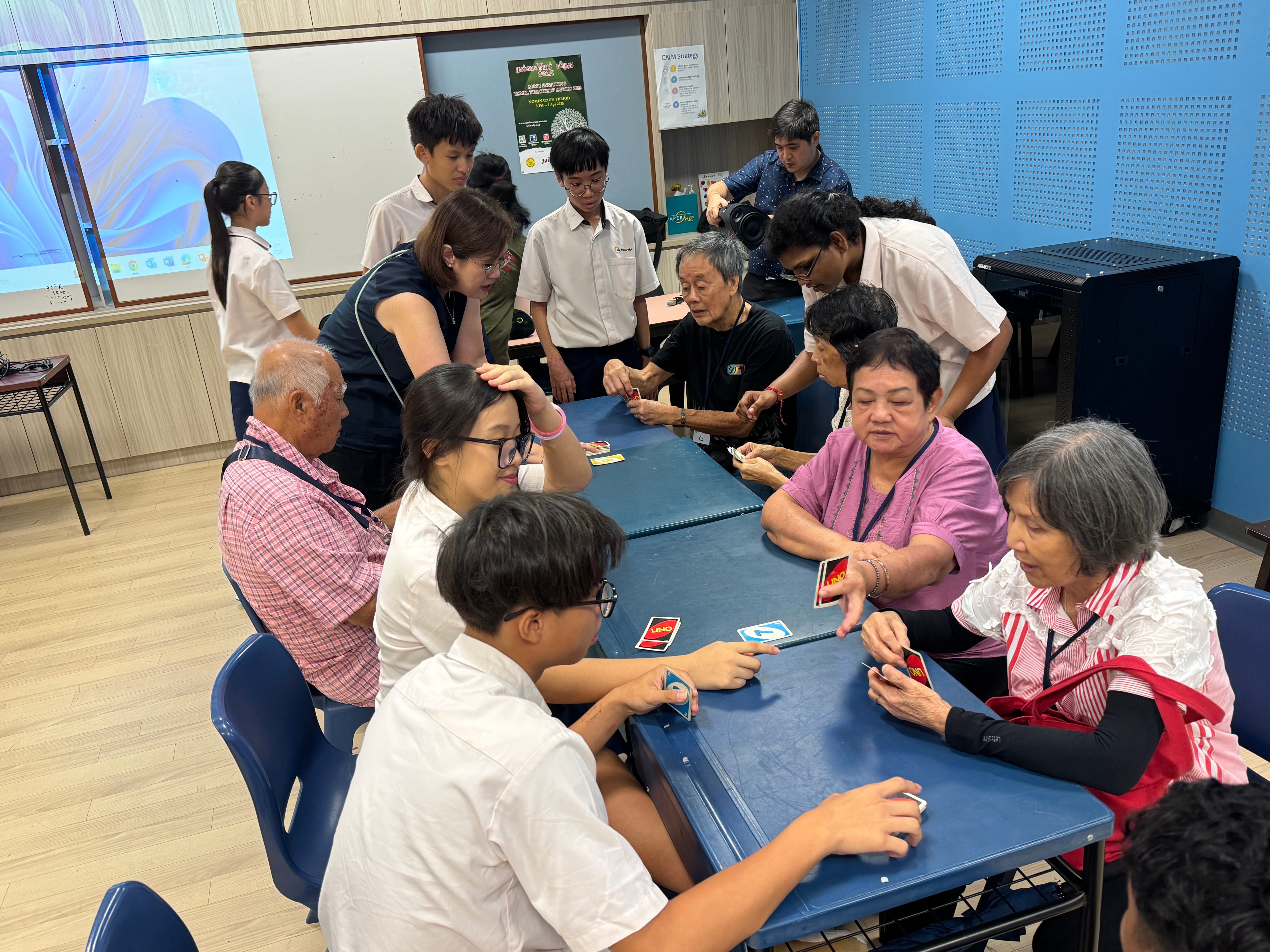 Students and seniors sit around blue tables playing UNO cards in a classroom while others watch.