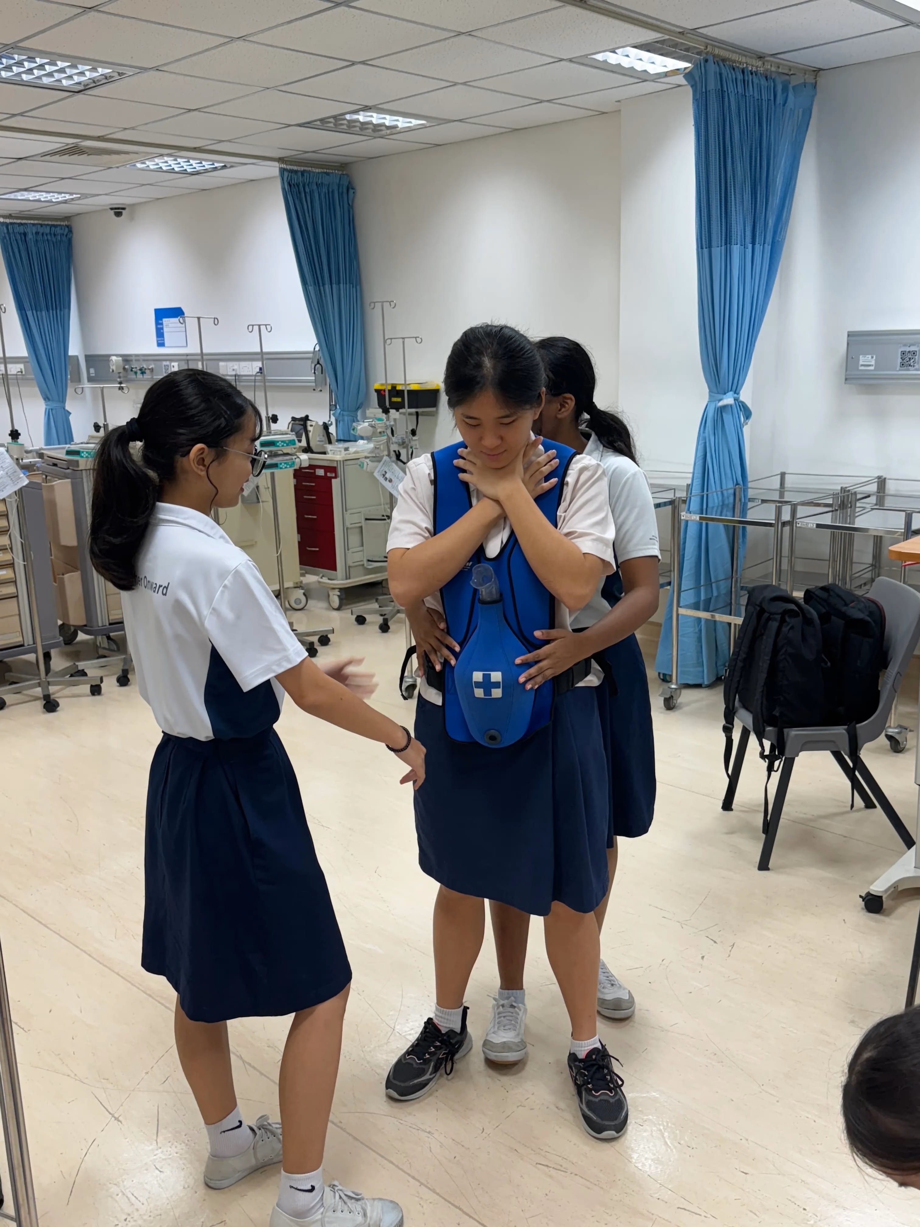 Three school girls in uniforms practice first aid in a medical training room, one wearing a blue CPR vest.